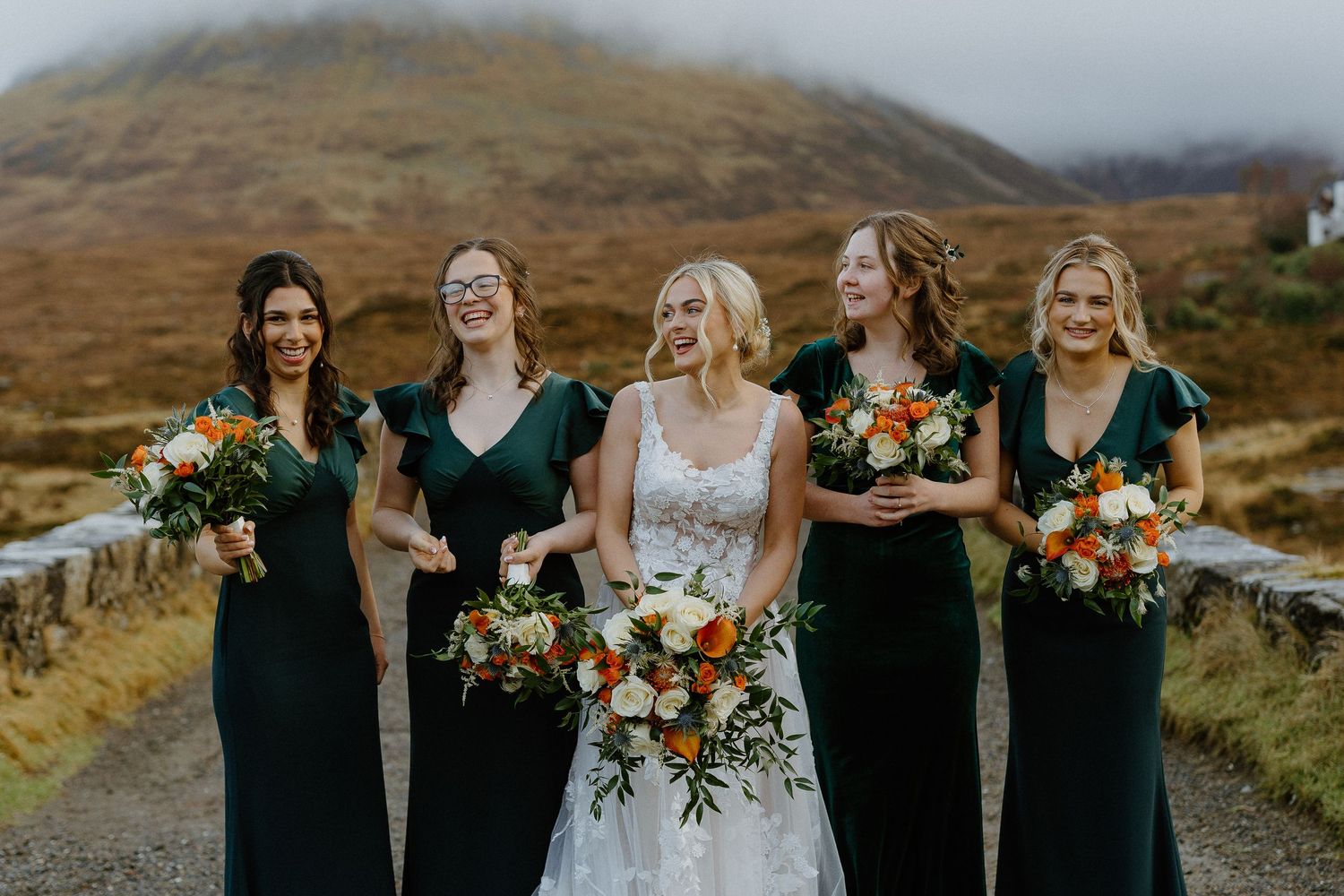 Bride in lace gown laughs with four bridesmaids in emerald green dresses holding bouquets outdoors.