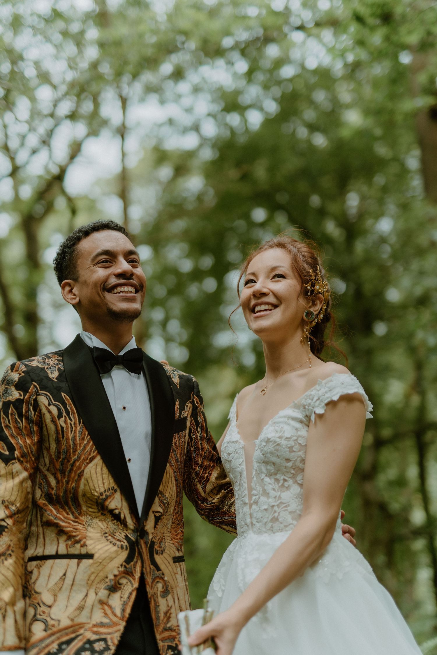 Smiling bride and groom in forest, groom in gold batik tuxedo, bride in lace wedding gown.