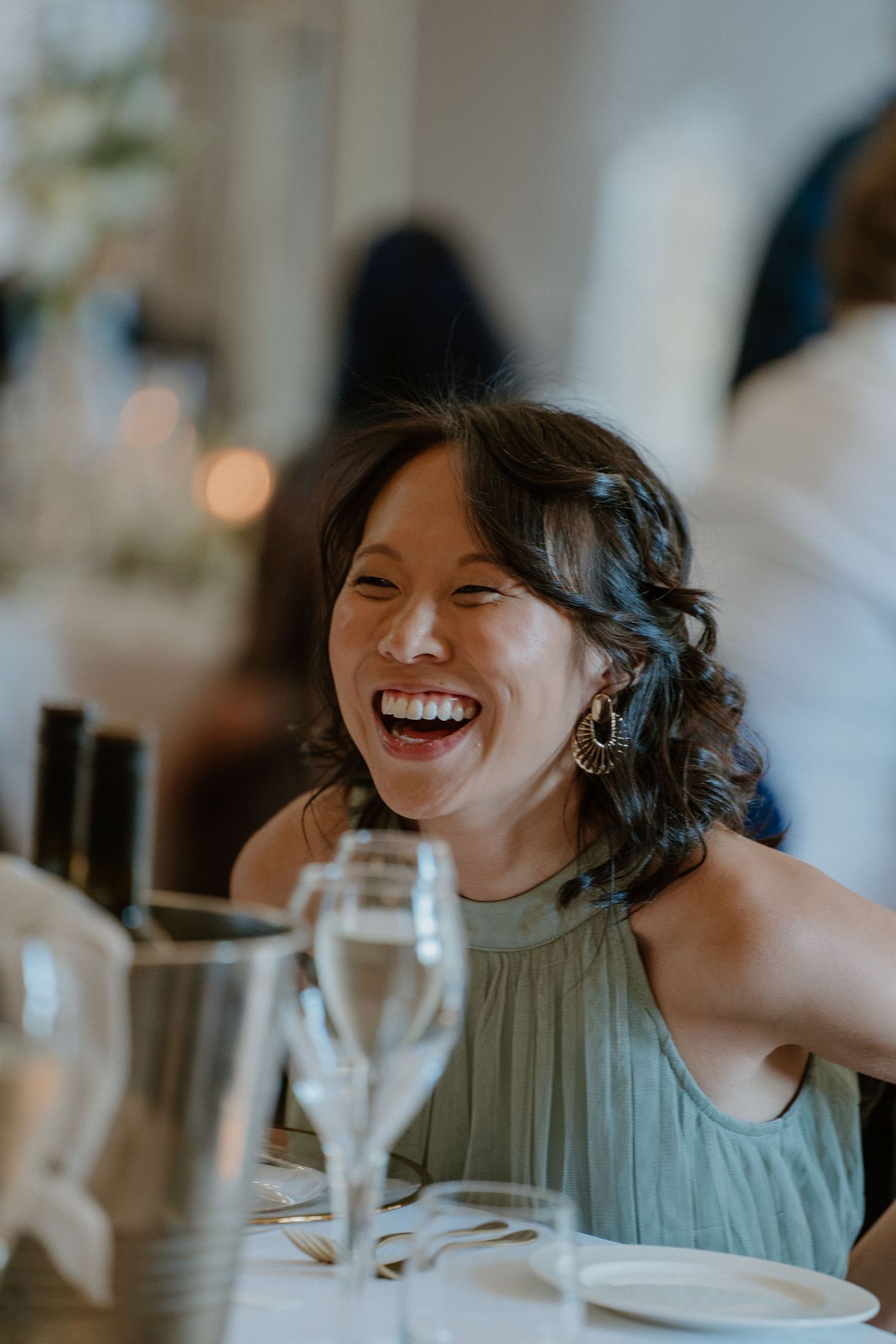 Wedding guest in sage green dress laughing joyfully at elegant reception dinner table.