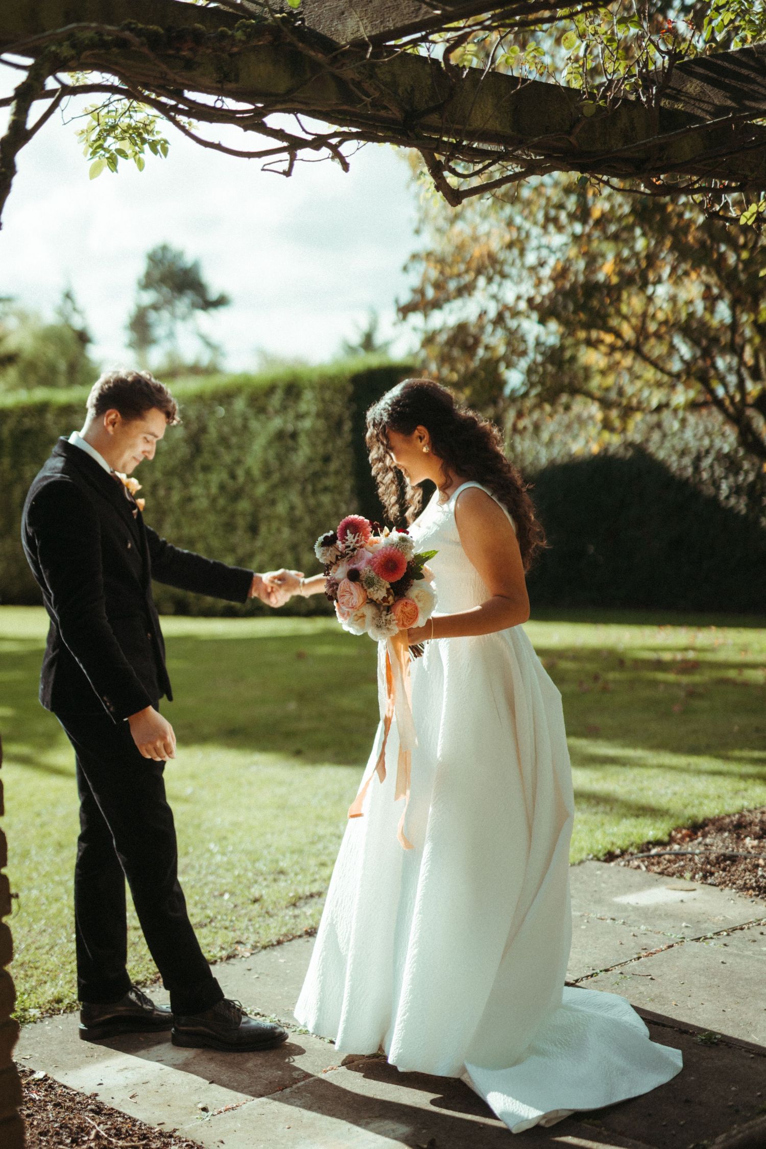 Bride and groom holding hands during romantic first look moment in sunlit garden under pergola.