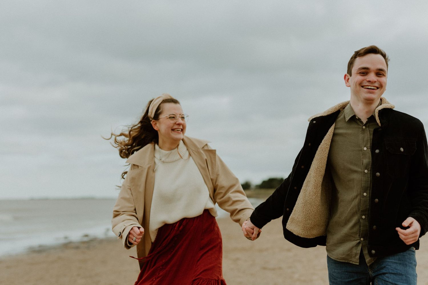 Happy couple holding hands and running along a sandy beach on a cloudy day.