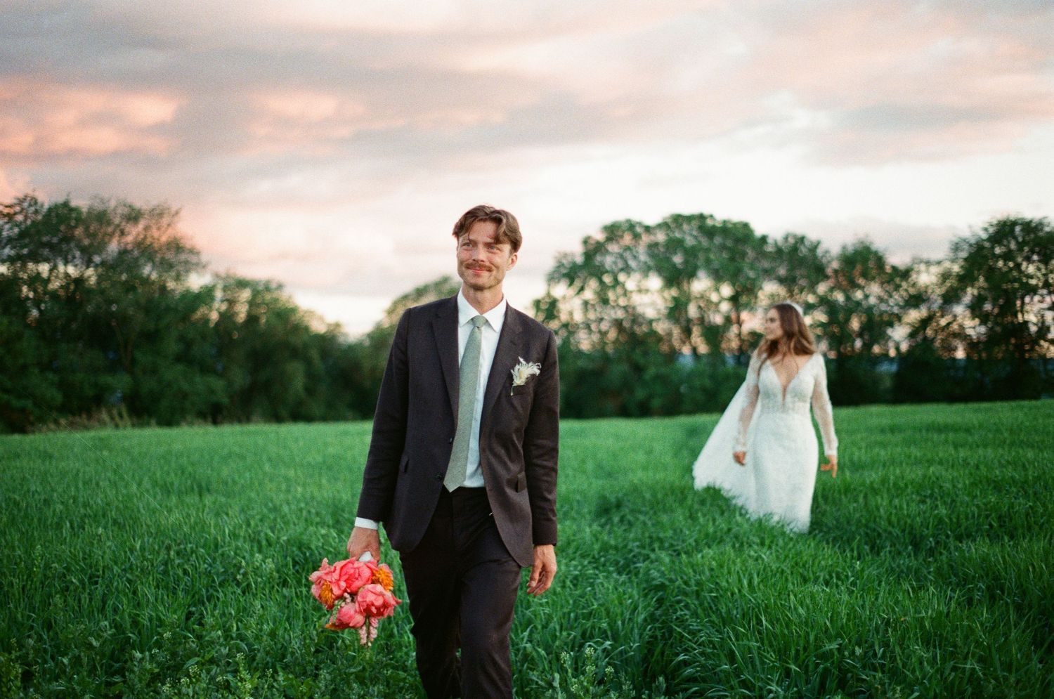 Groom holding pink bouquet walks through lush green field as bride in white gown follows at golden hour sunset.