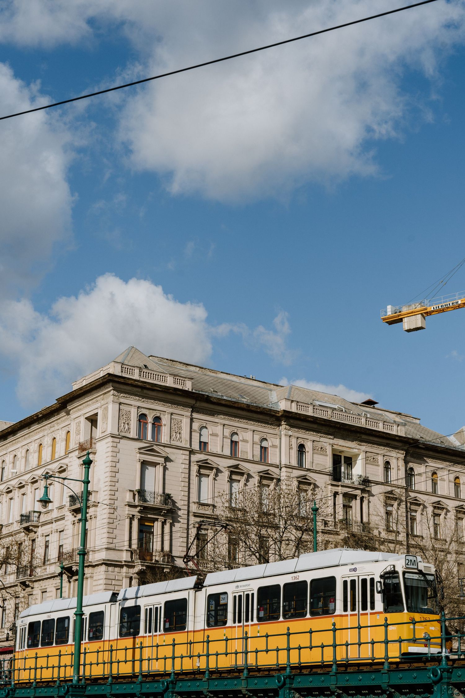 Yellow tram passes by historic stone building in Budapest with construction crane visible against blue cloudy sky.