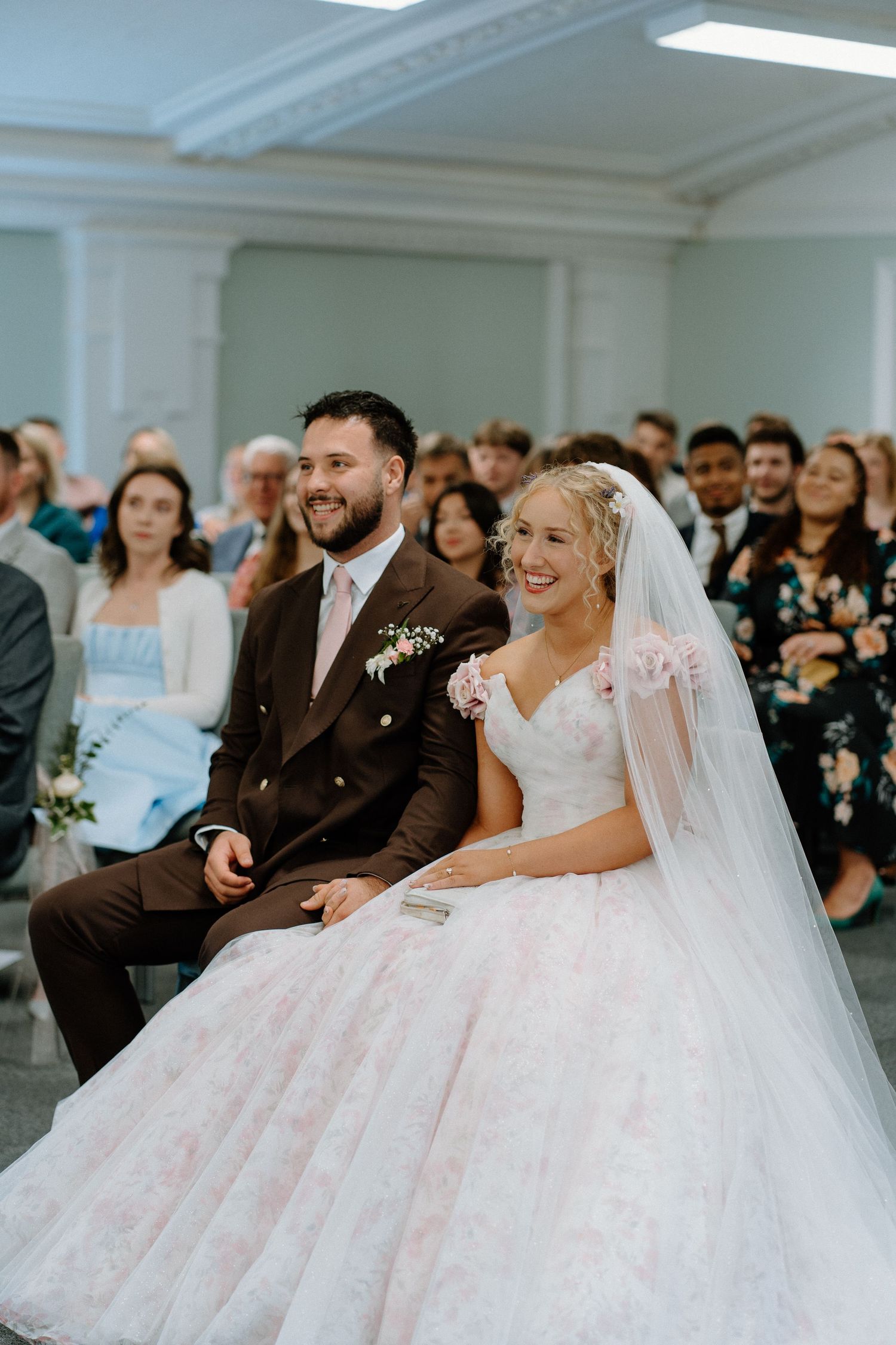 A joyful bride in a tulle ballgown and veil sits next to her groom during their wedding ceremony indoors.