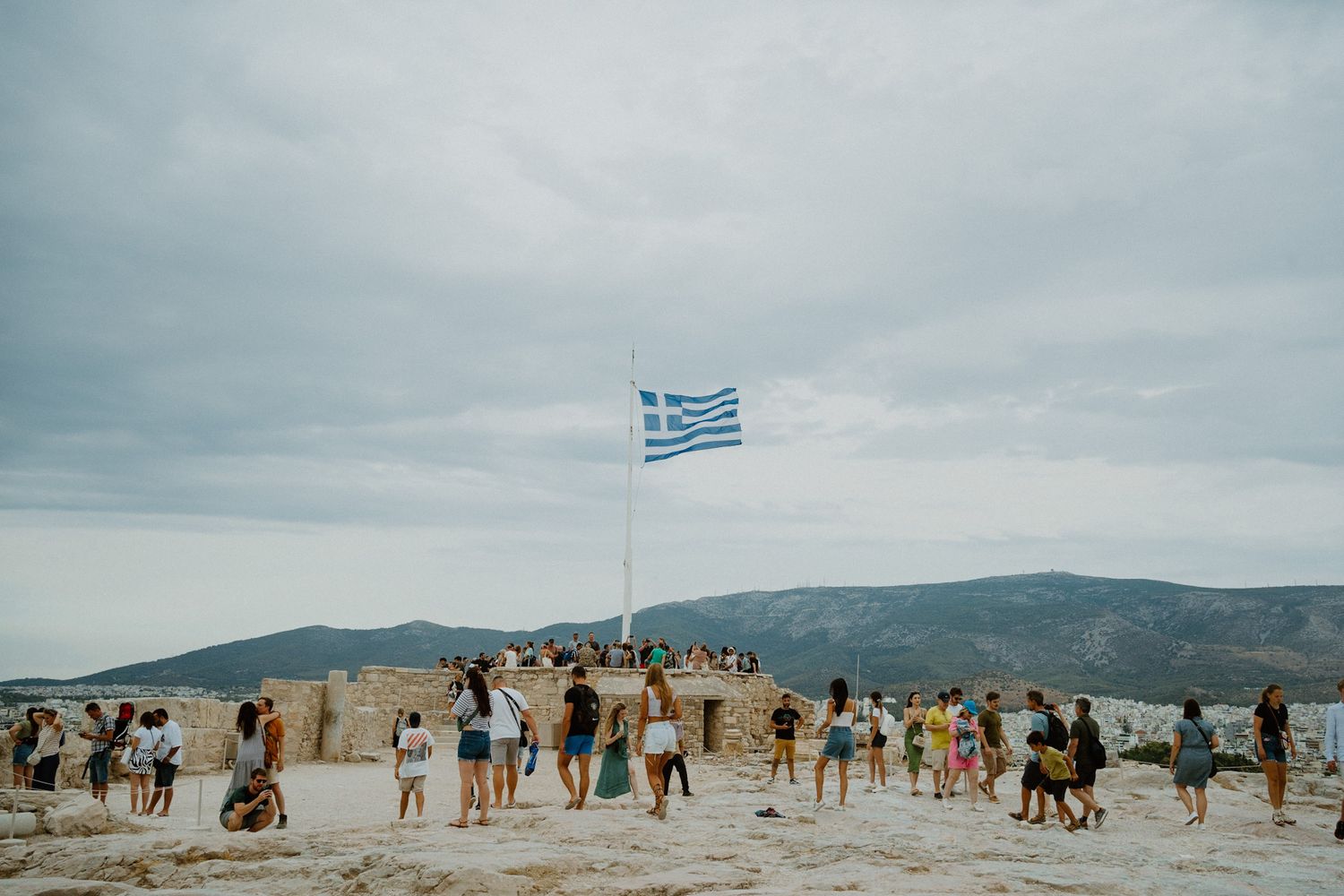Large group of tourists gather at hilltop viewpoint with Greek flag waving against cloudy sky and mountain backdrop.