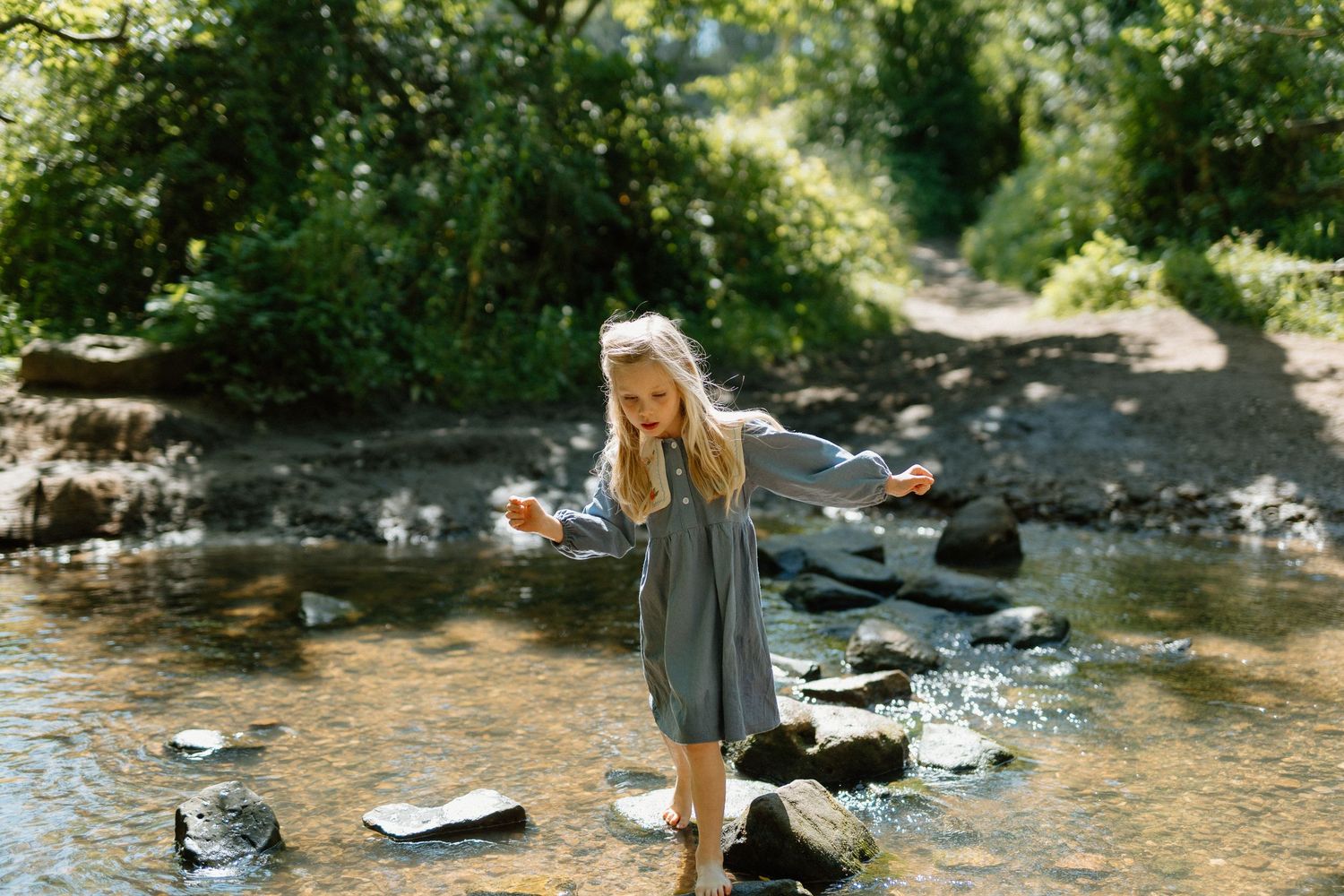 Young blonde girl in grey dress balancing on rocks in a shallow sunlit woodland stream.