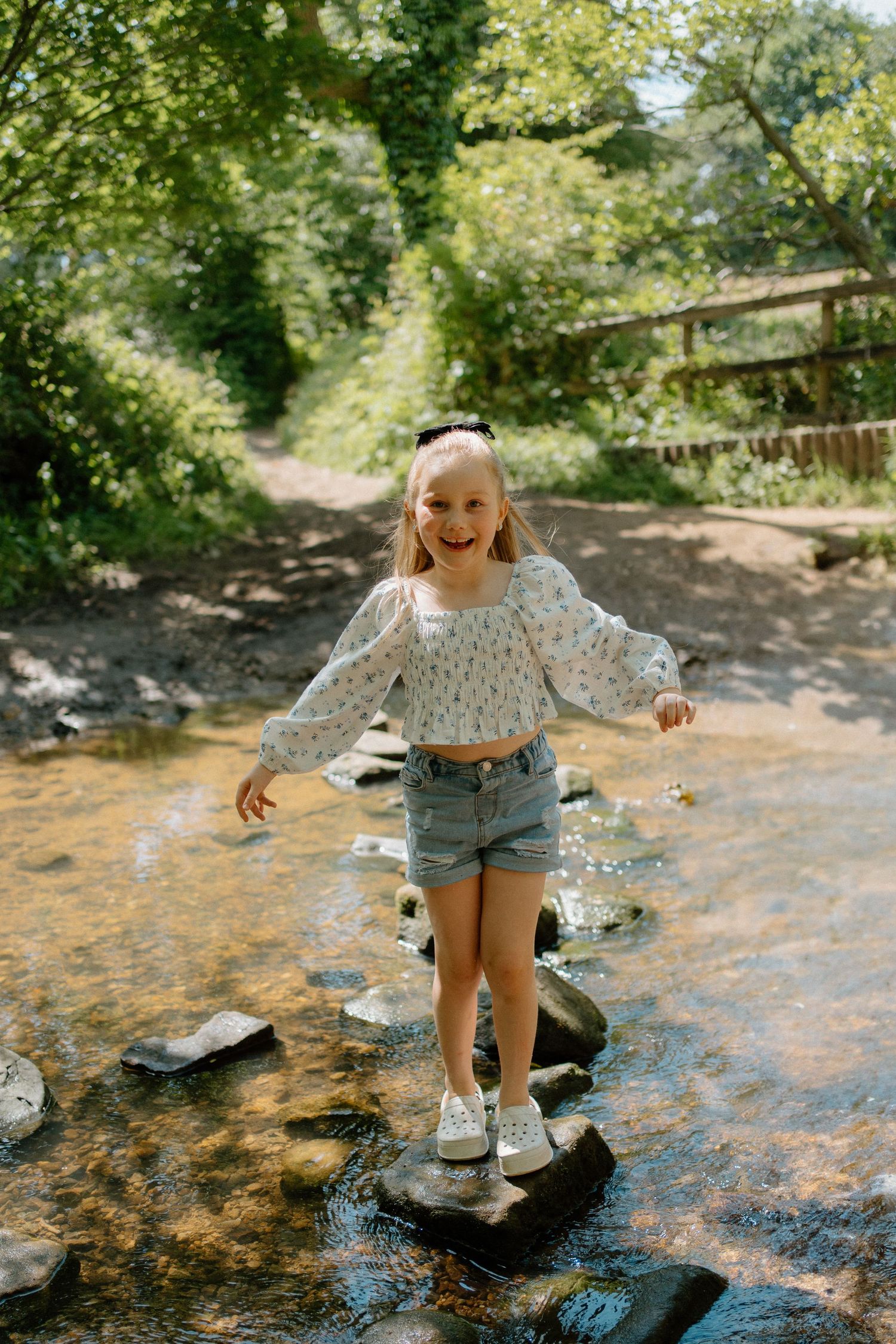A smiling girl in denim shorts crosses a shallow creek by stepping on rocks in a forest.