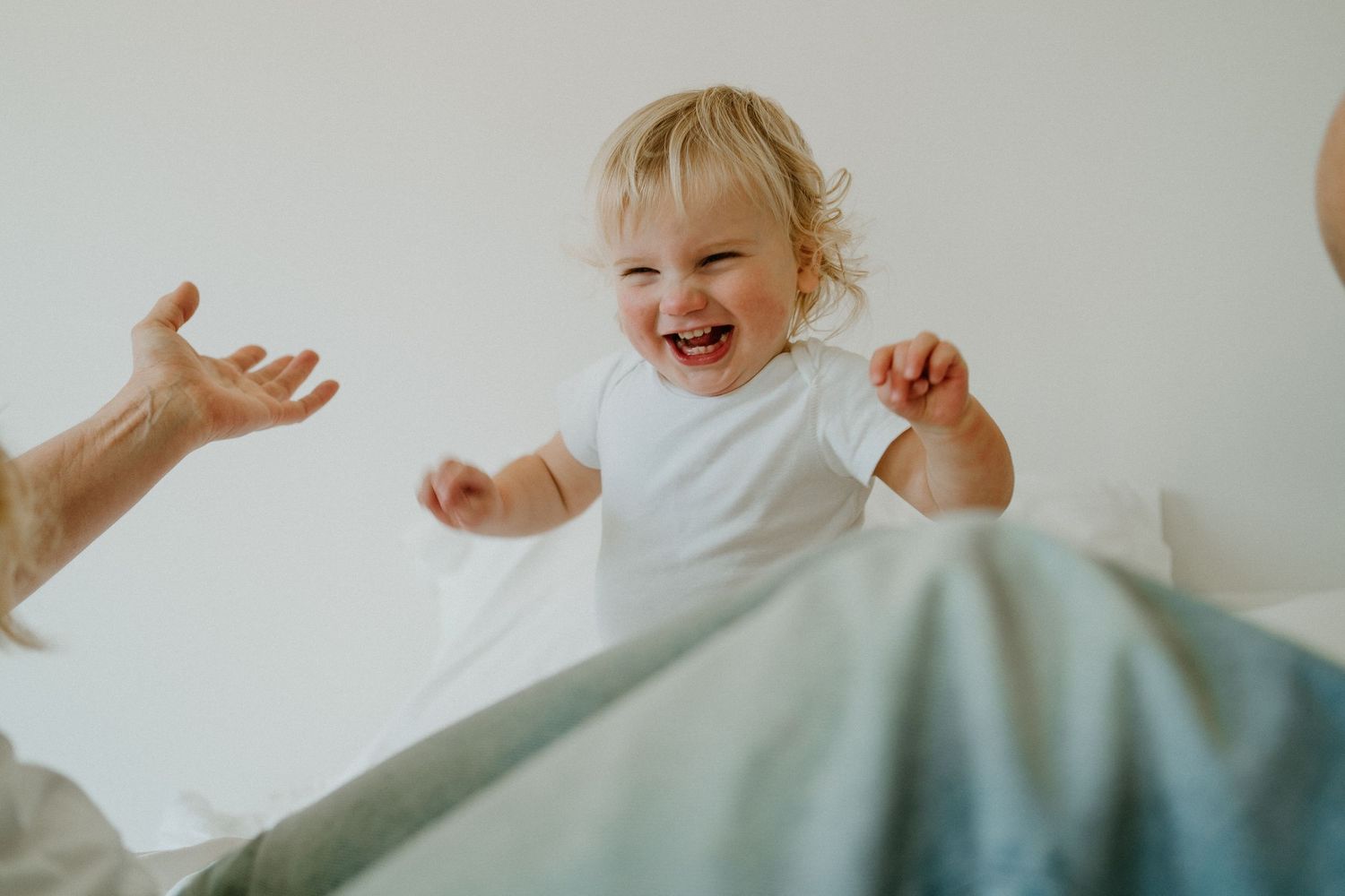 Joyful baby in white onesie laughing and reaching out during playtime on a bed.