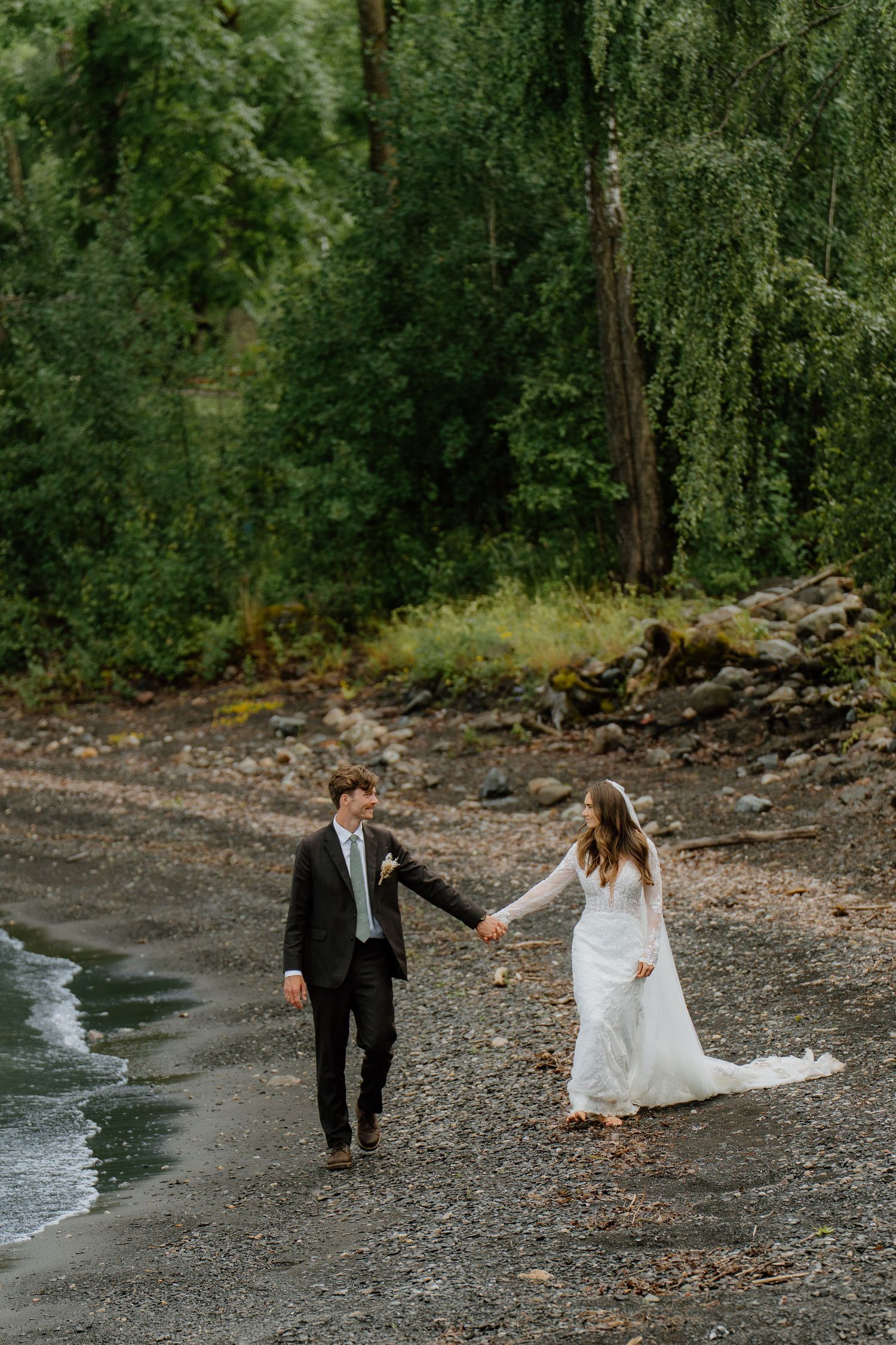 Bride and groom holding hands walking along rocky beach shoreline near lush forest.