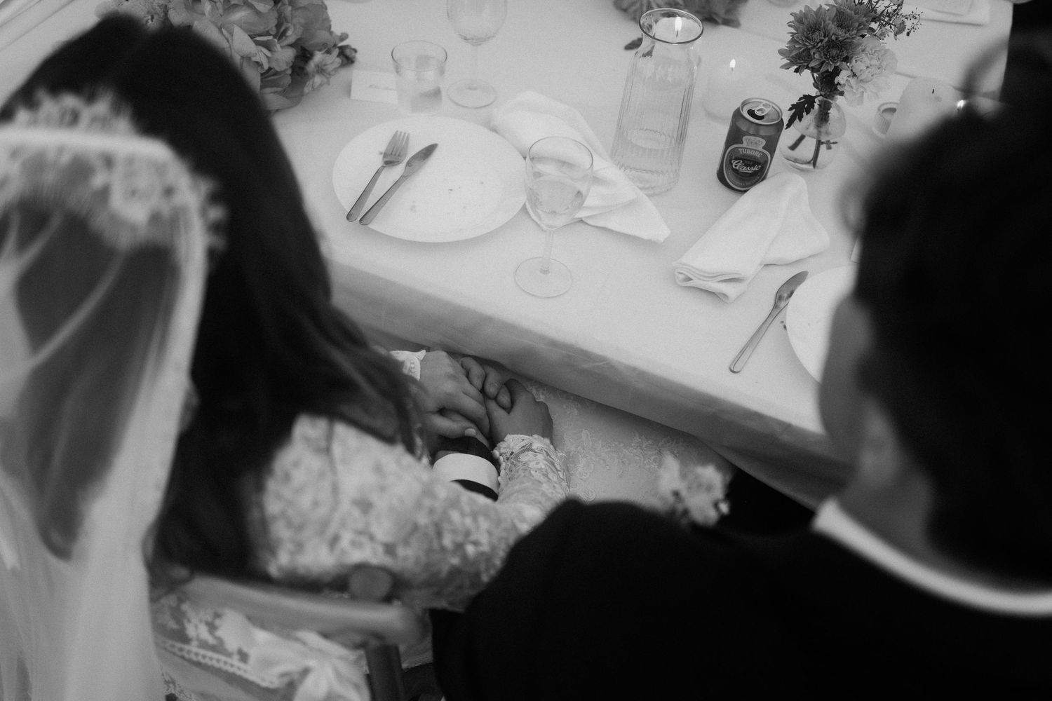 Black and white photo of bride and groom holding hands under the wedding reception table.