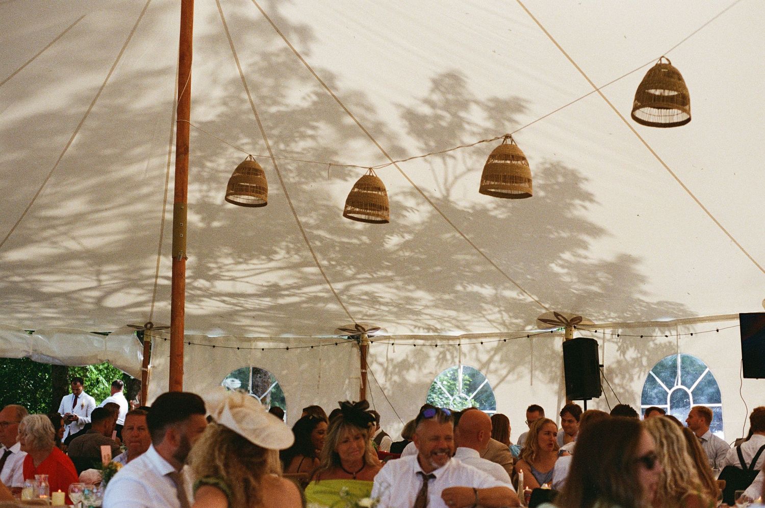Wedding guests dining under a marquee tent with wicker pendant lights and fairy string lights overhead.