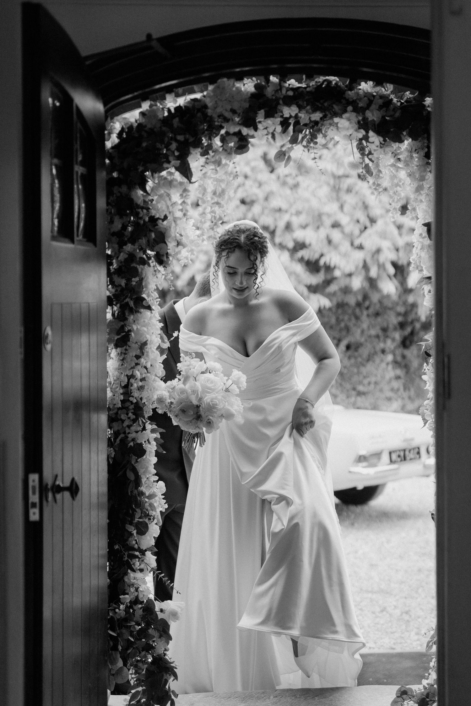 Bride in off-shoulder gown holding bouquet framed by floral doorway arch.