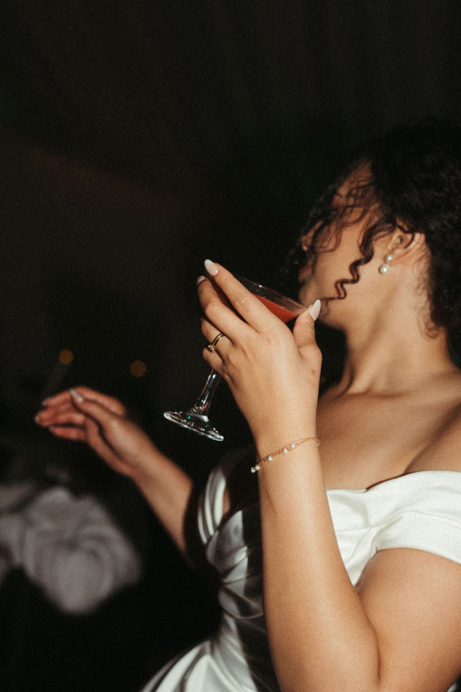 Bride in white off-shoulder gown sipping red wine from elegant cocktail glass at wedding reception.
