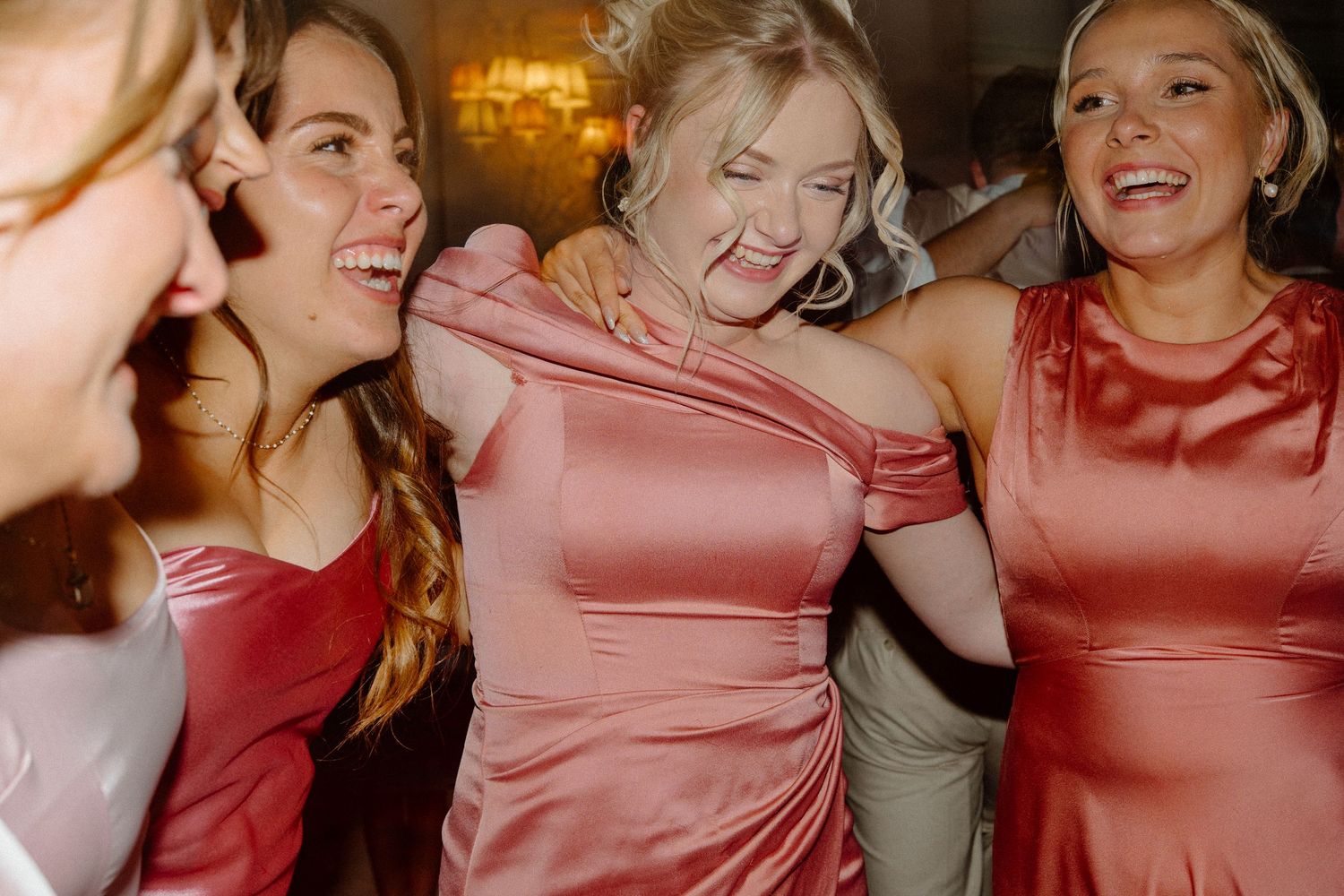 Bridesmaids in pink satin dresses laughing and dancing together at a wedding reception.