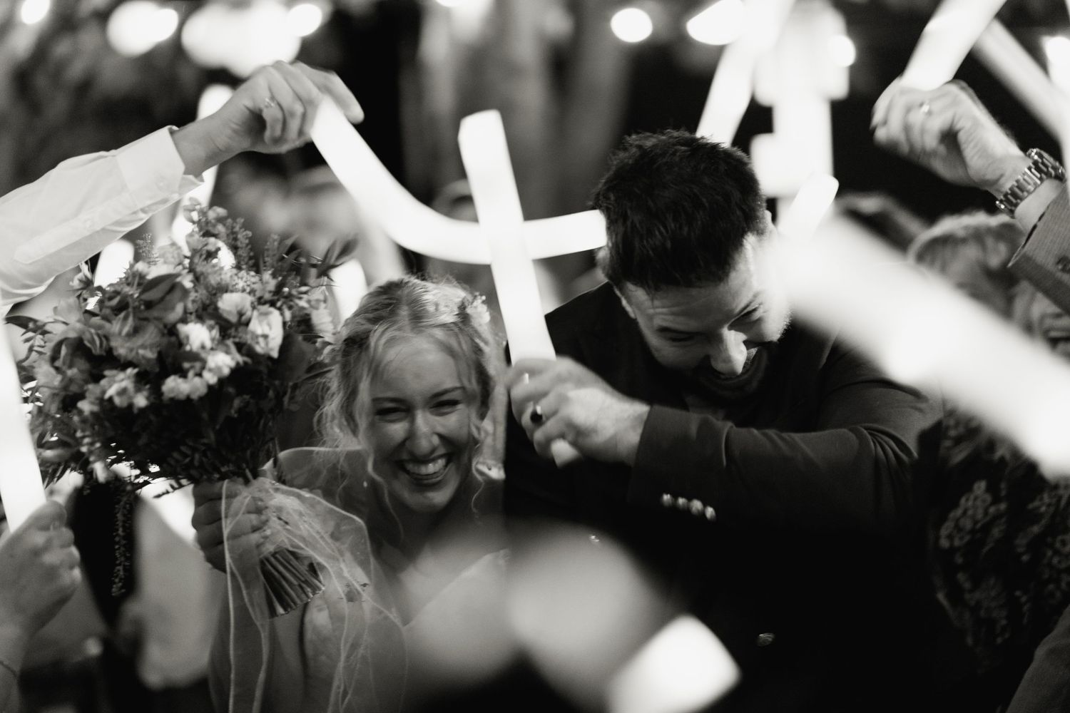 Black and white photo of laughing bride and groom walking through guests waving glowsticks.