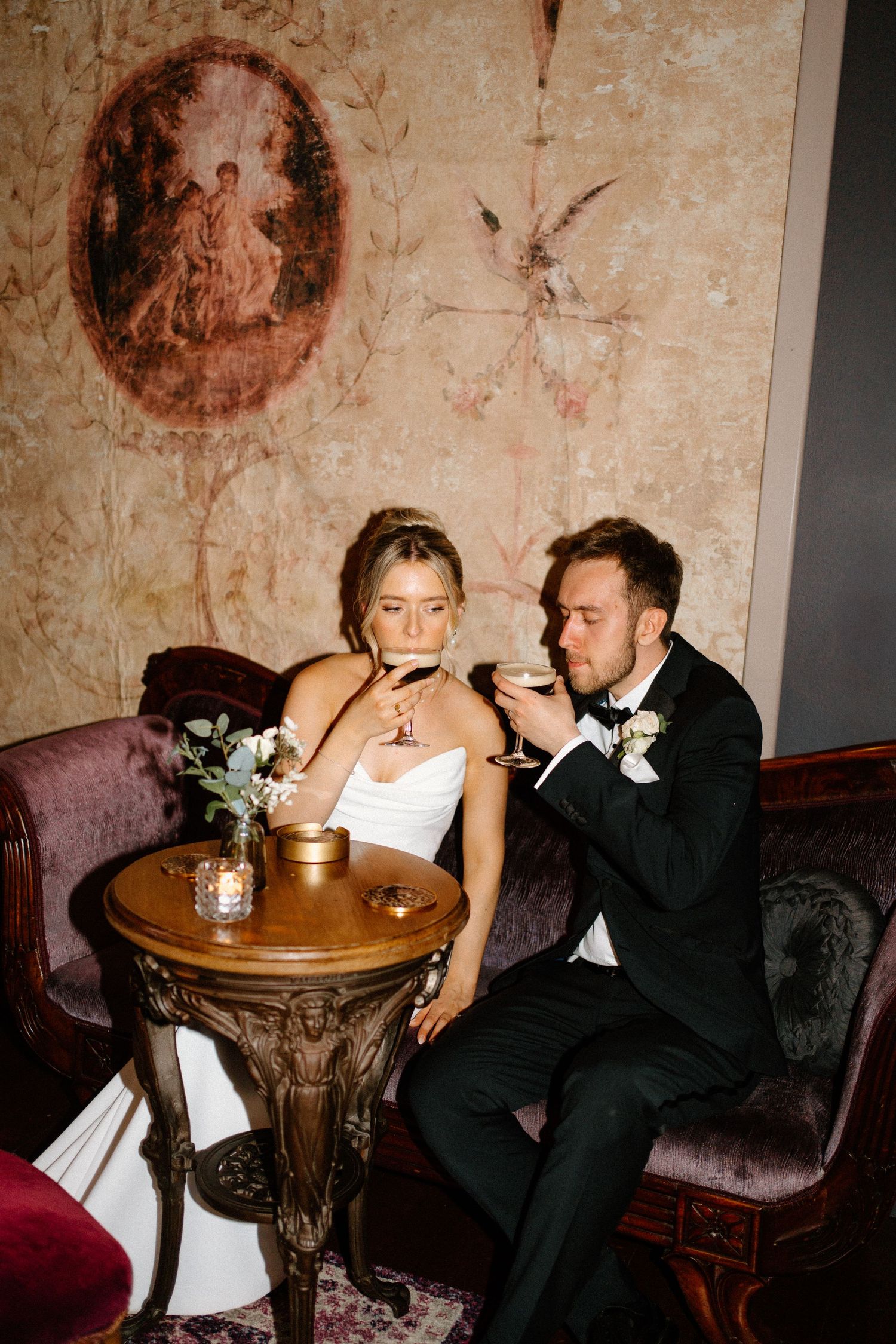 Bride and groom sit together on velvet sofa sipping drinks beside ornate table in luxurious decorated venue.