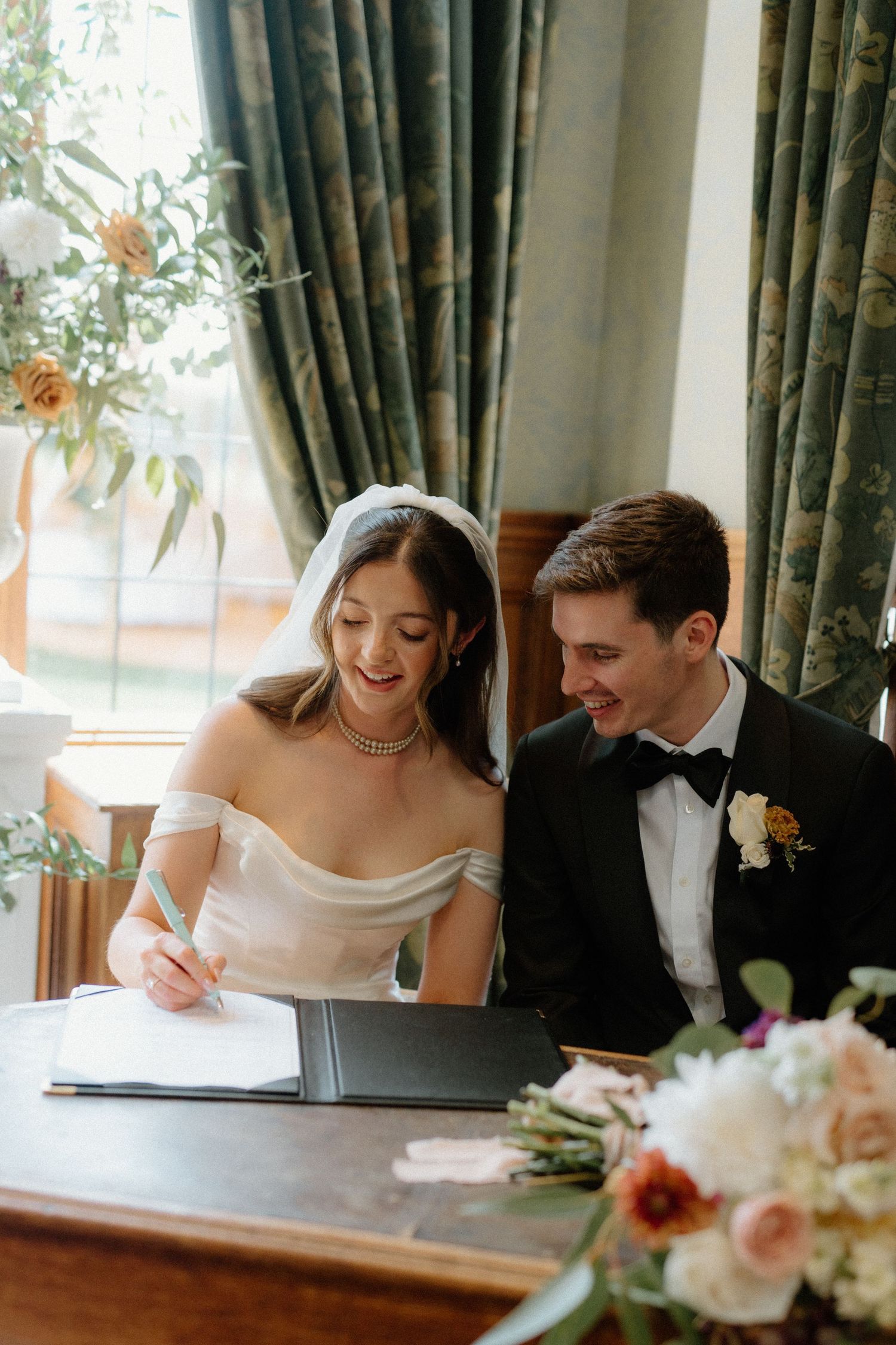 Bride in white gown signs wedding register while groom in black tuxedo watches and smiles.
