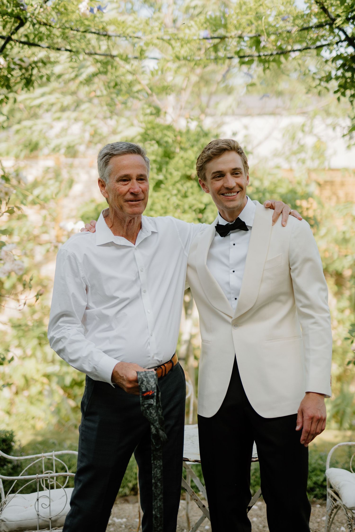 Groom in white tuxedo poses with older man in white shirt at elegant outdoor garden wedding.