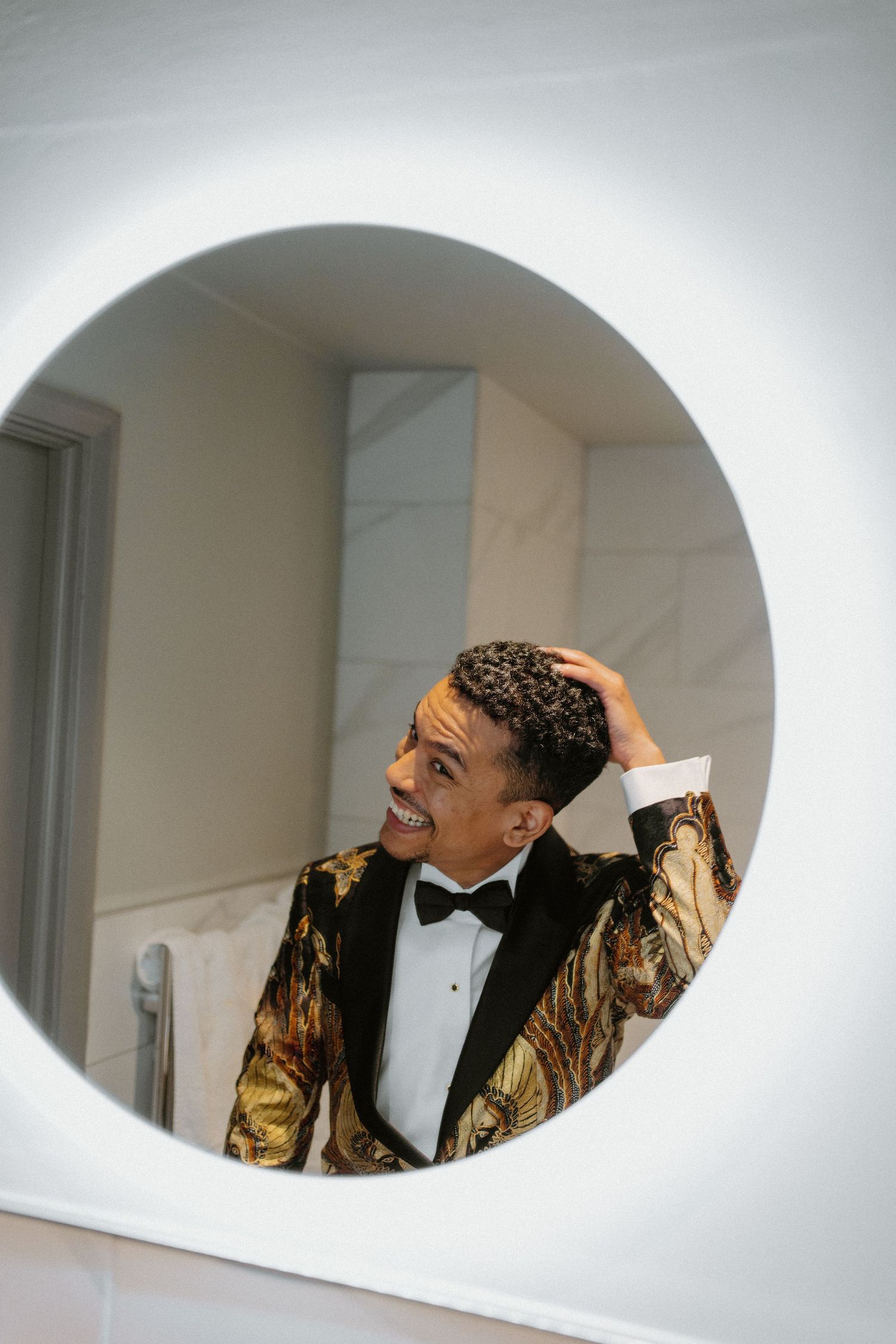 Groom in gold patterned tuxedo jacket smiles at reflection in round lit mirror.