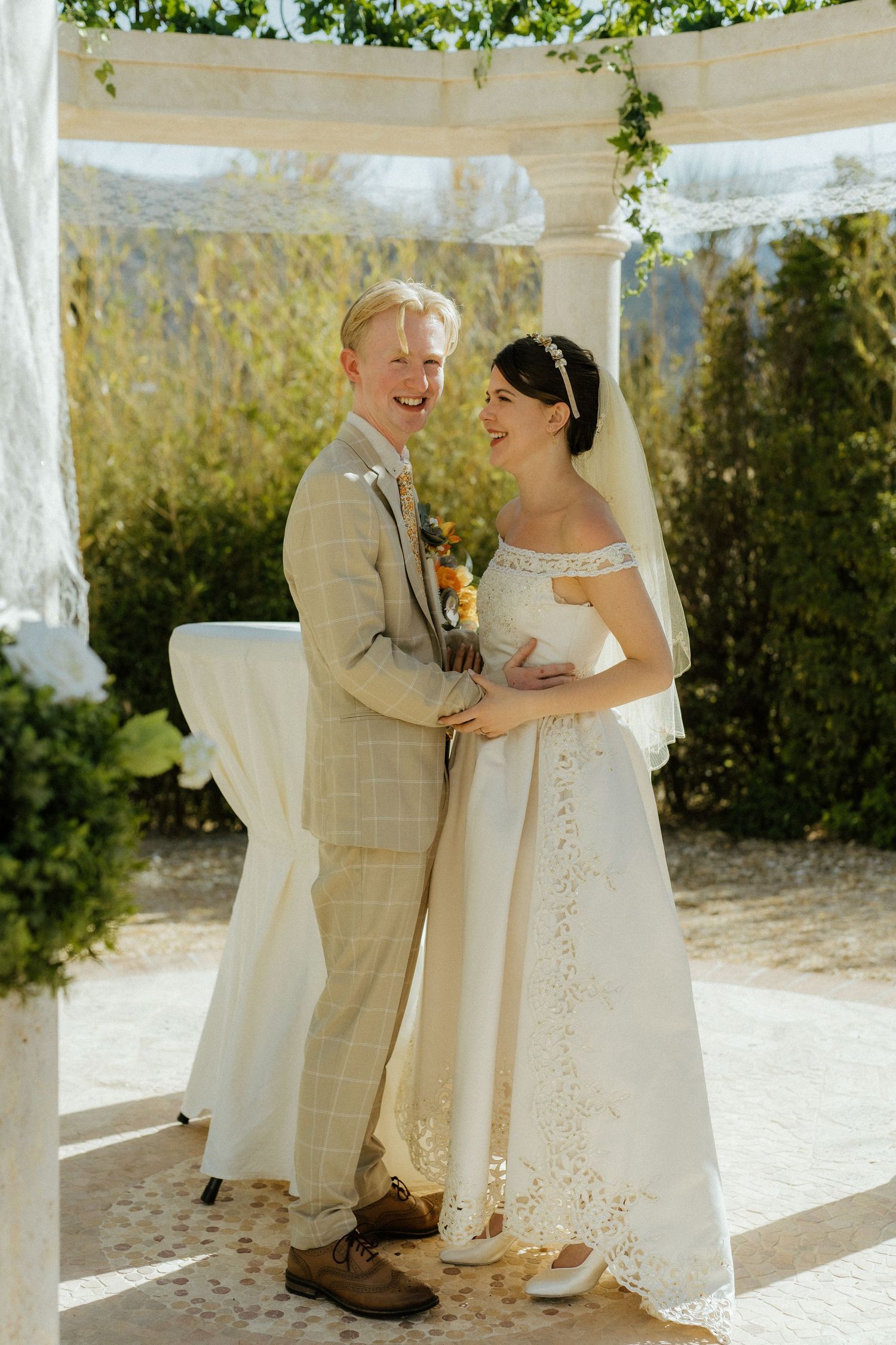 Happy bride and groom laughing together under white floral arch after outdoor wedding ceremony.
