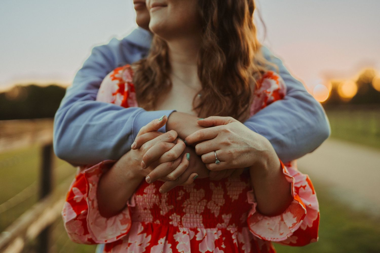 Close-up of hands resting on a red floral dress against a sunset background in a grassy field.
