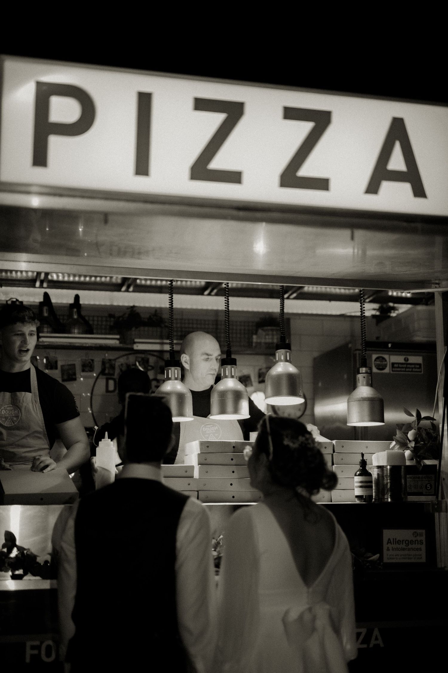 Black and white photo of a busy pizza counter with workers and stacked pizza boxes under a glowing PIZZA sign.