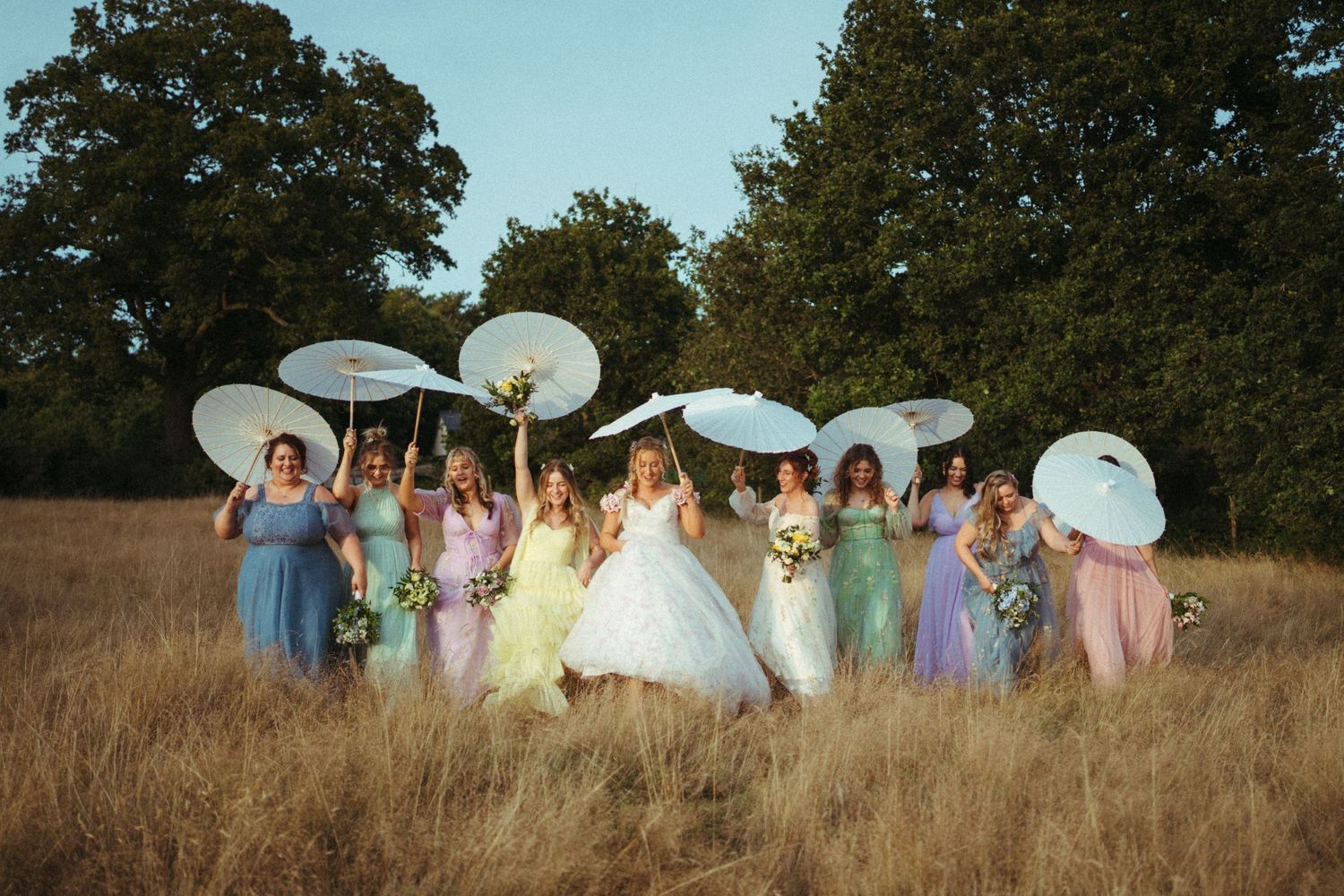 Wedding party with white parasols poses together in a golden field at sunset surrounded by green trees.