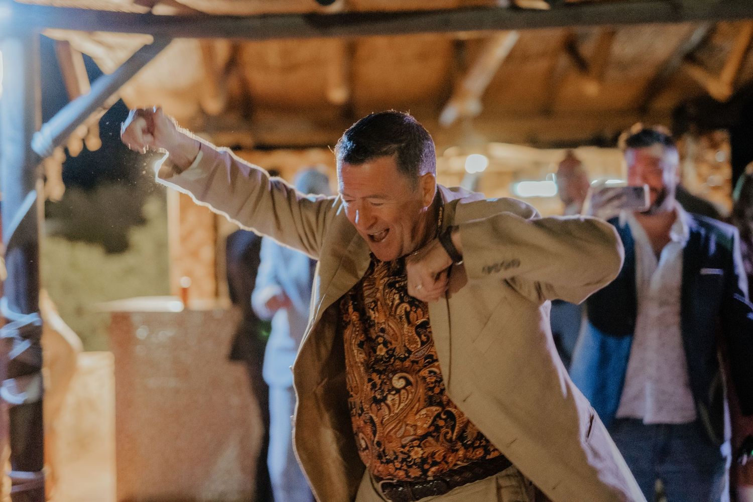 Joyful wedding guest dancing energetically in tan blazer at rustic barn reception.