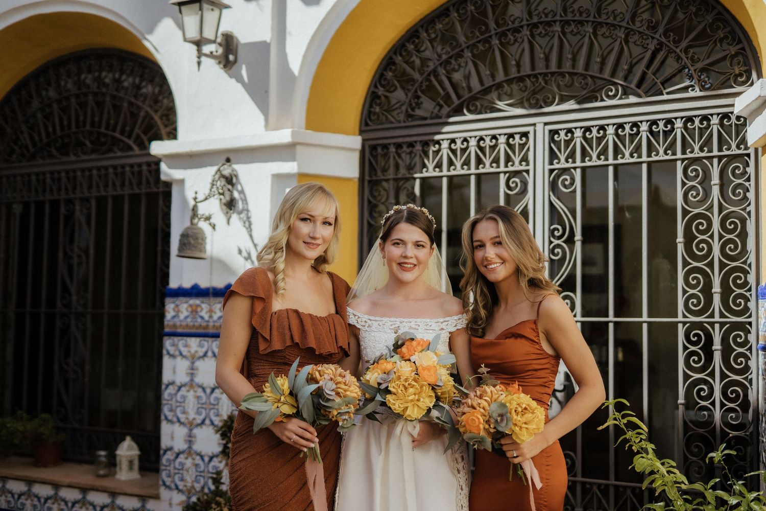 Bride in white gown posing with two bridesmaids in rust-colored dresses holding yellow bouquets.