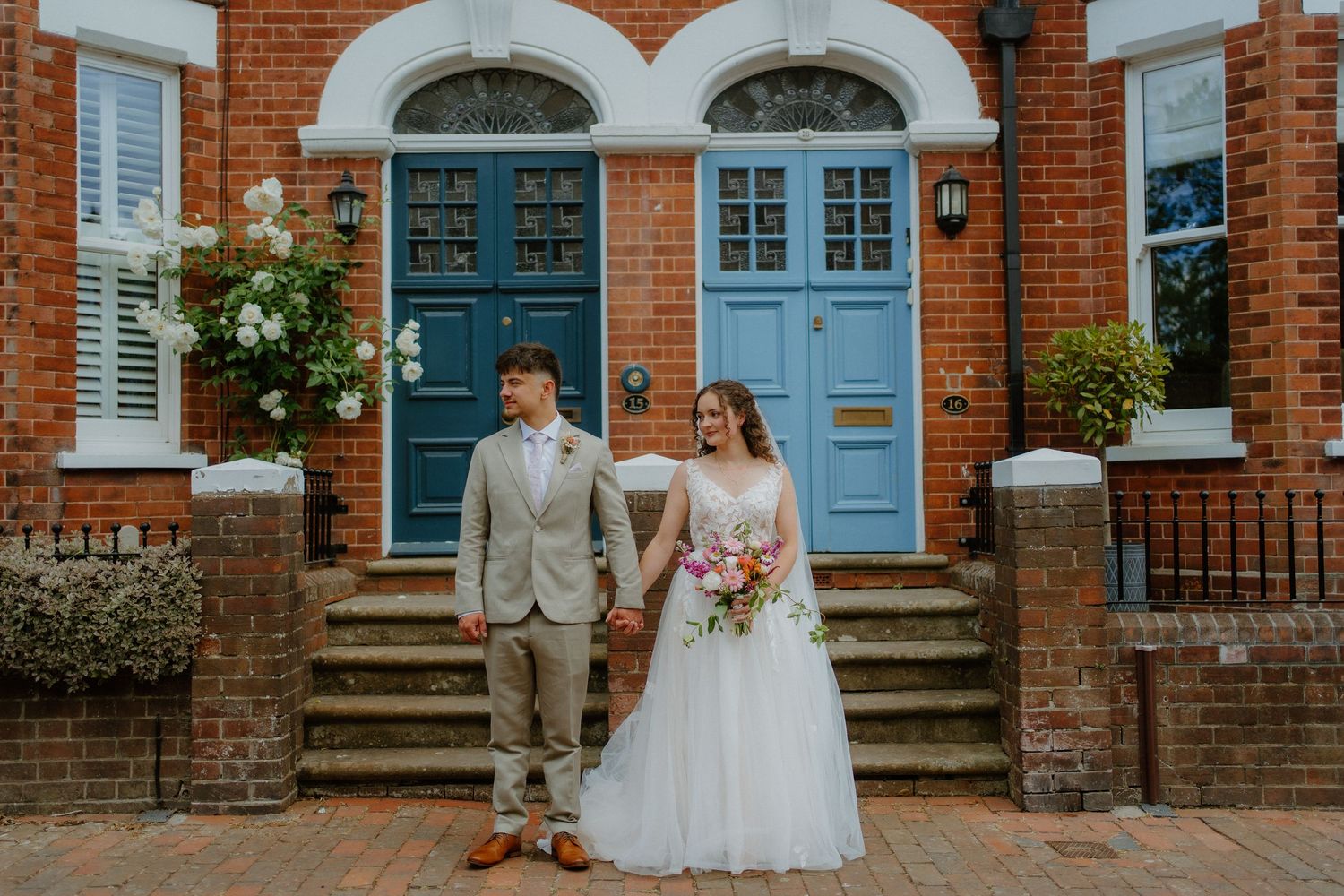 Bride and groom holding hands on brick steps before blue doors in an elegant outdoor wedding portrait.