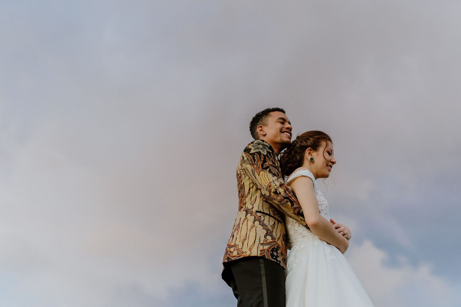 Couple in wedding attire standing back-to-back against a cloudy sky backdrop.