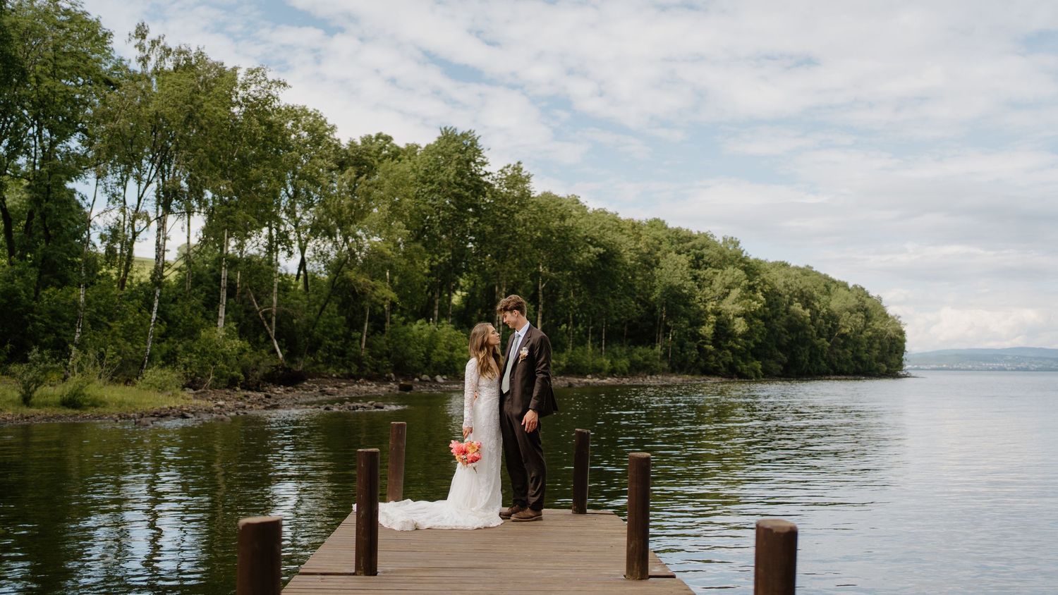 Bride and groom share romantic kiss on wooden dock overlooking calm lake with lush trees.