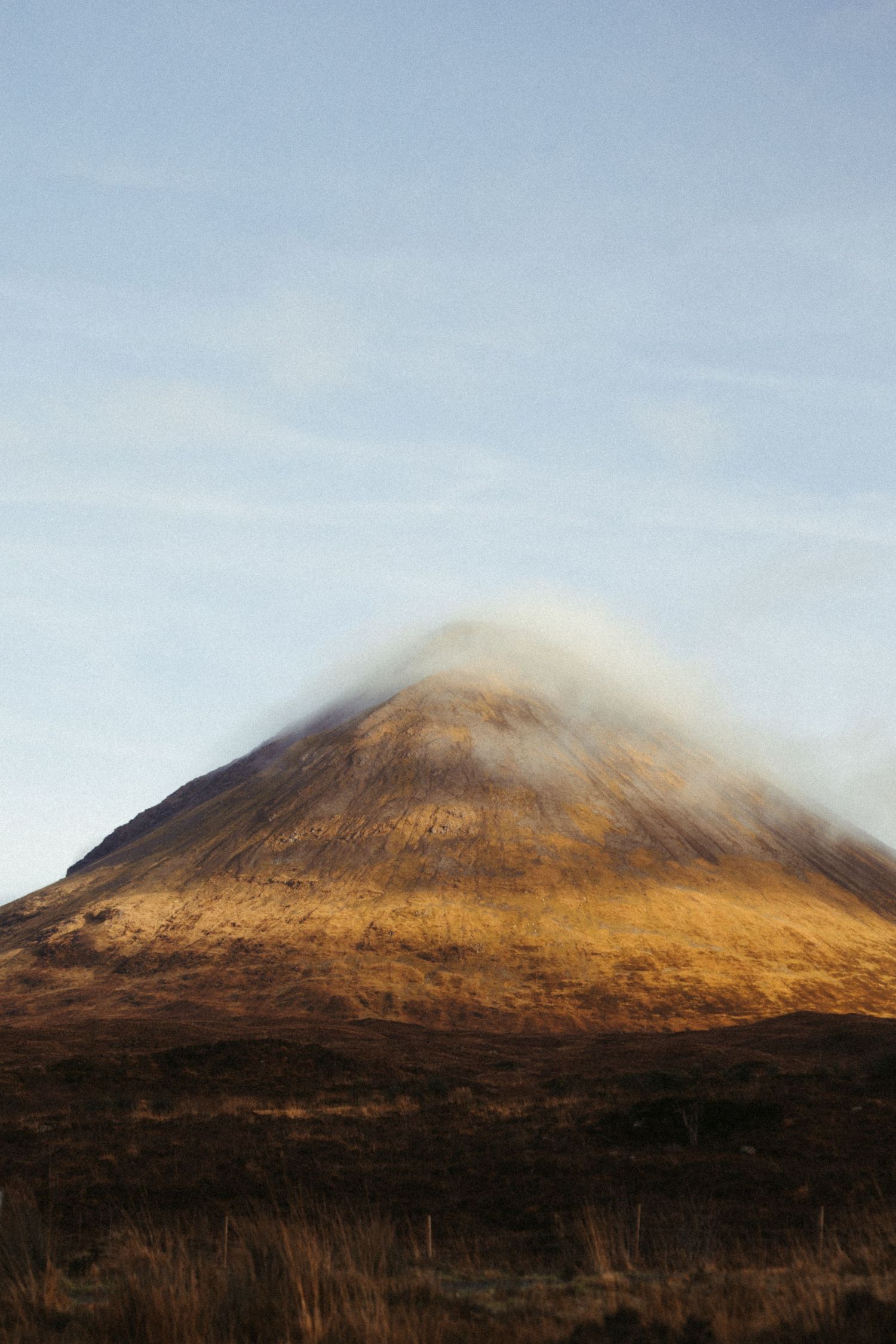 Golden sunlight illuminates a dramatic cone-shaped Scottish Highland mountain peak partially shrouded in low cloud.