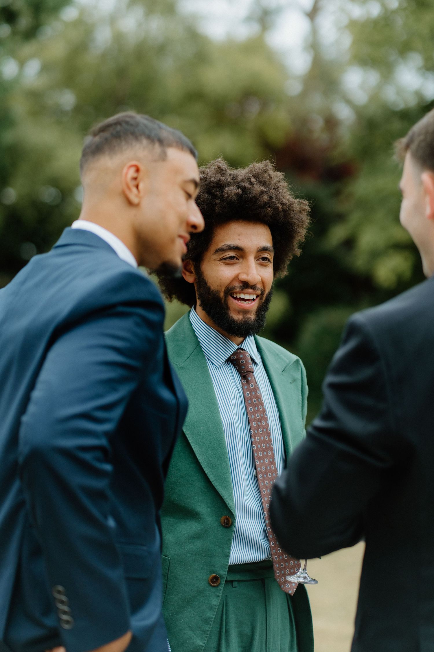 Smiling male wedding guest in green suit chats with friends during outdoor wedding reception.