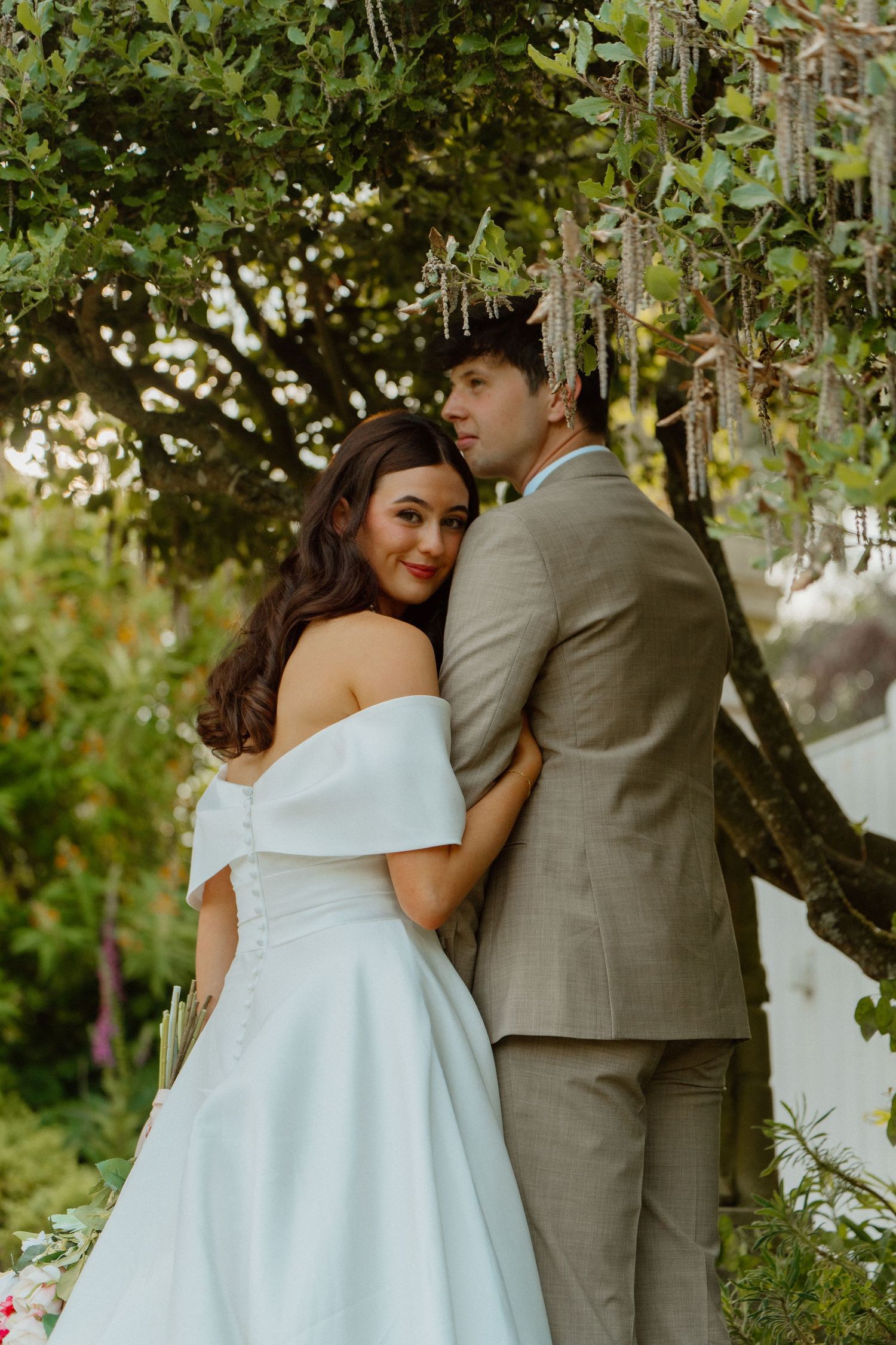 Bride in an off-shoulder white gown embracing groom in a tan suit under a decorated tree at their garden wedding.