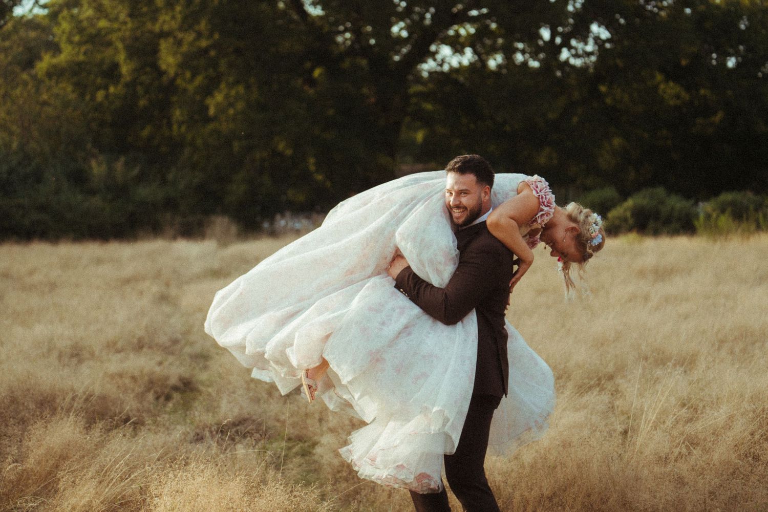 Groom joyfully lifts bride in flowing white dress while spinning in a sunlit golden meadow.