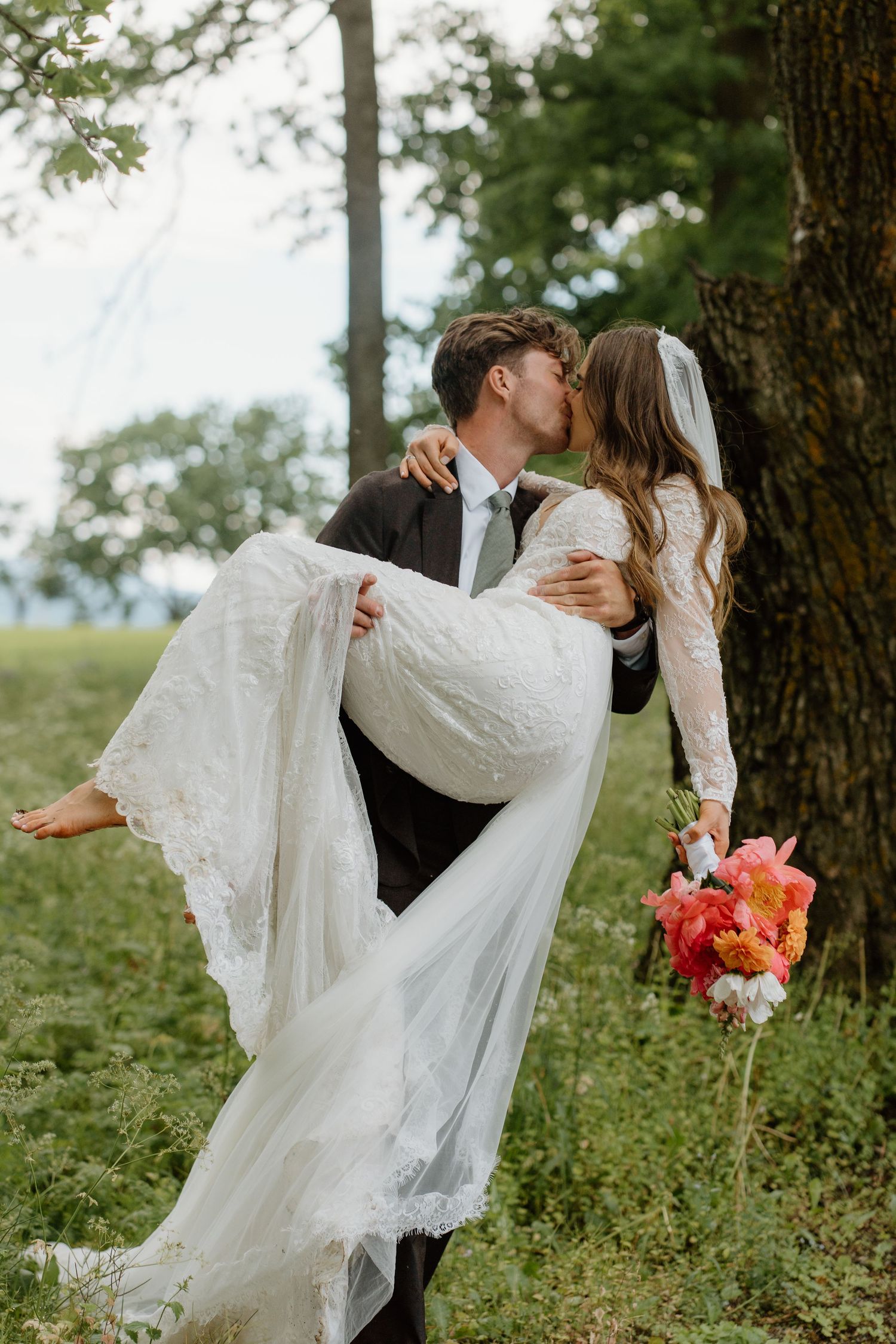 Groom carrying bride in white lace dress while kissing near tree in green meadow.