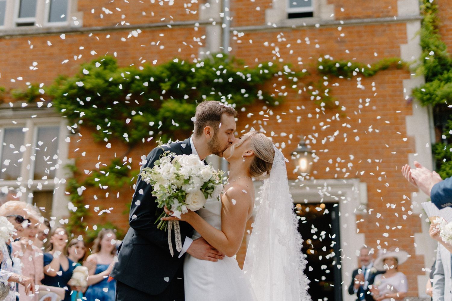 Bride and groom kissing surrounded by a shower of white confetti petals outside a brick manor house.