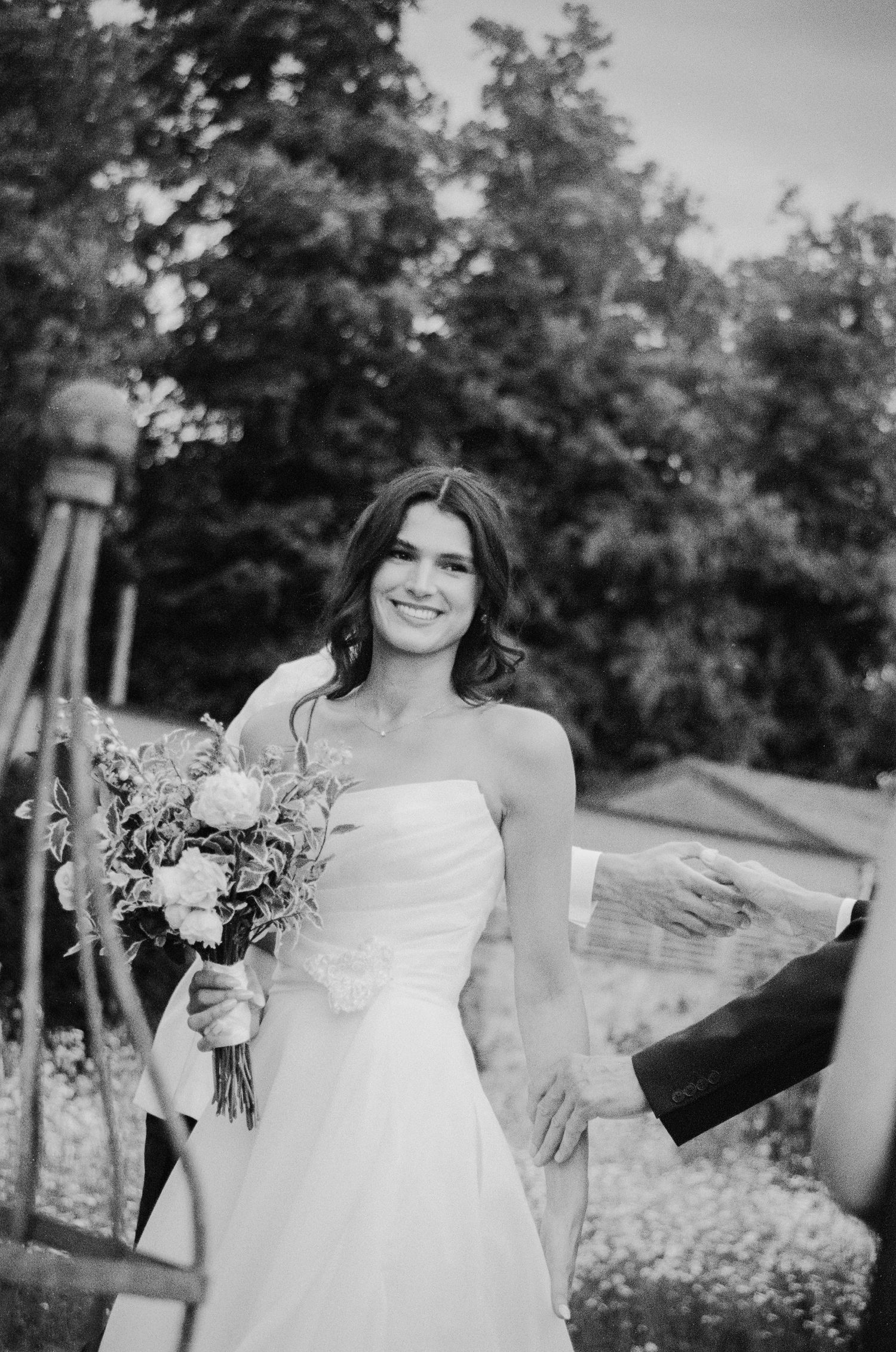 Smiling bride in strapless white gown holding floral bouquet in black and white photo.