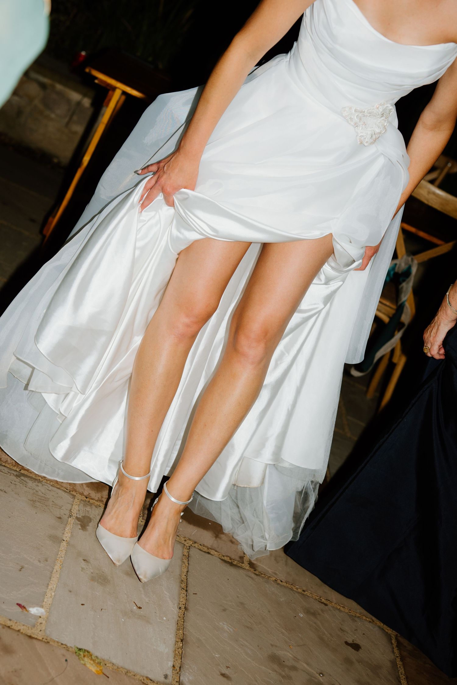Close-up of bride lifting white wedding gown revealing heels on stone patio.