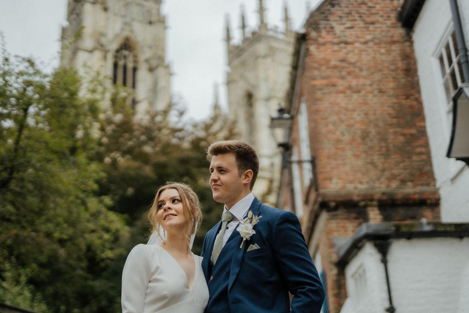 Elegant couple in wedding attire standing near a historic brick church tower surrounded by lush greenery.