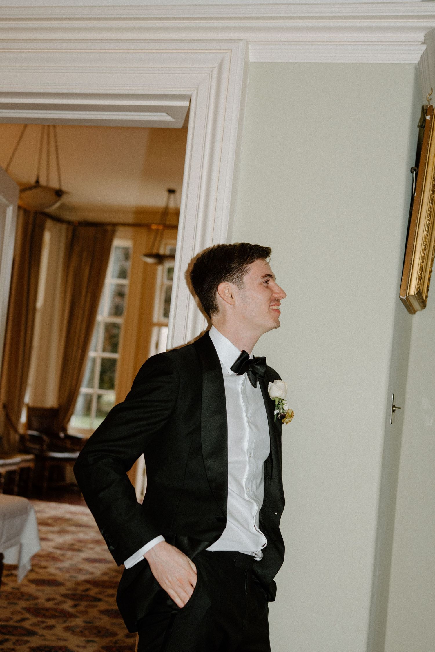 Dapper groom in black tuxedo with bow tie and boutonniere leaning against an interior wall.