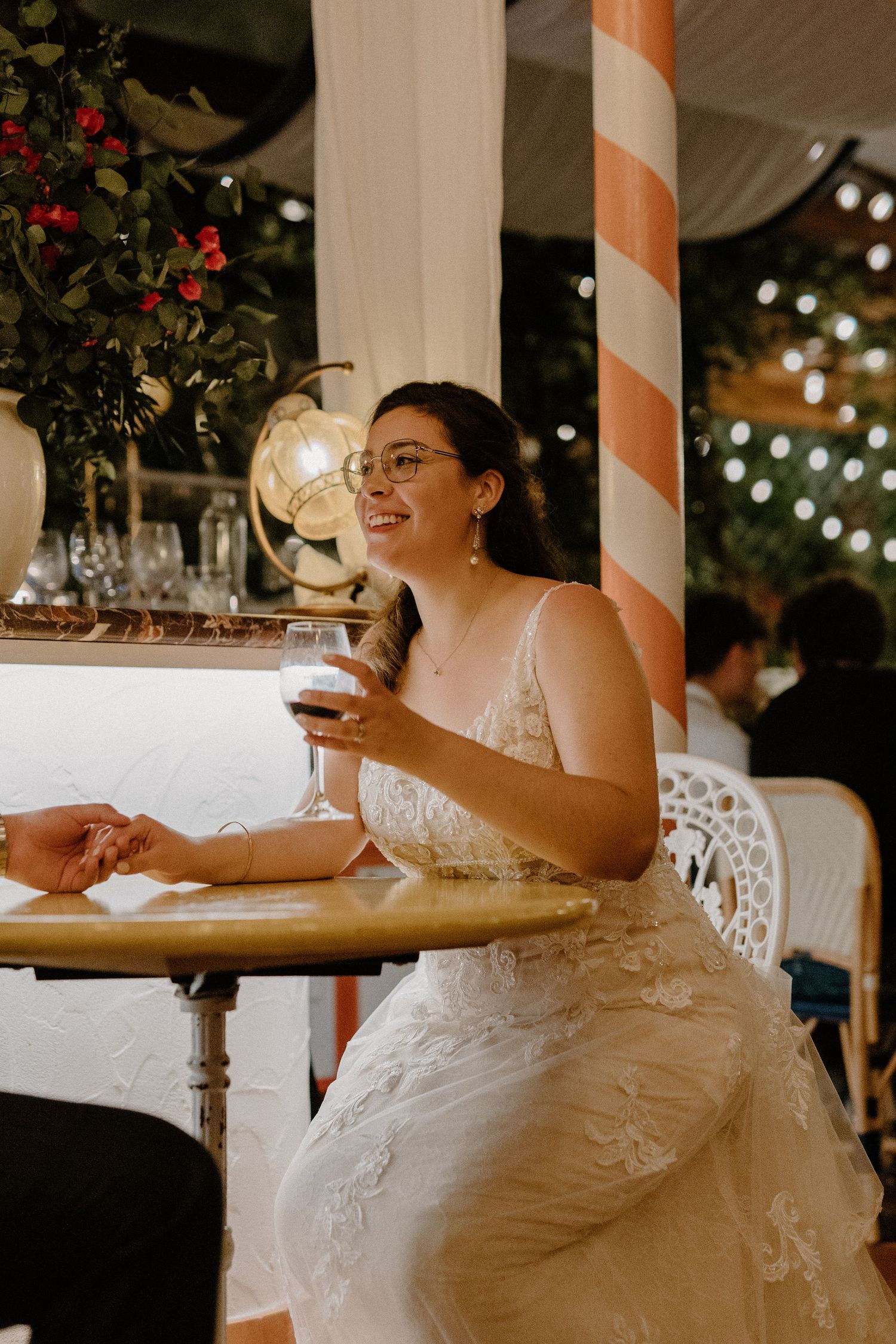 Smiling bride in lace gown holding a wine glass while seated at a decorated wedding venue table.