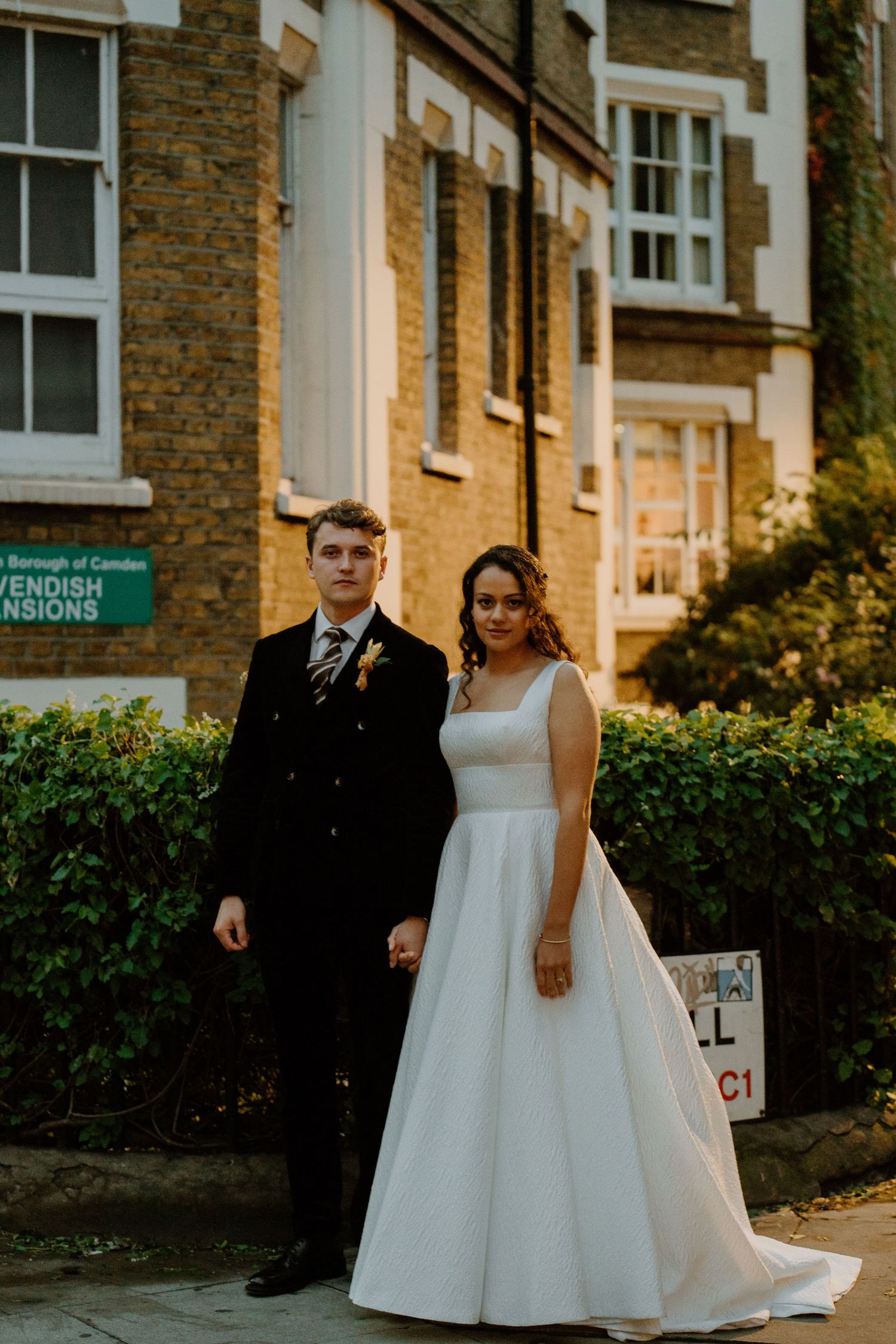 Elegant bride and groom posing hand in hand outside a brick building in London.