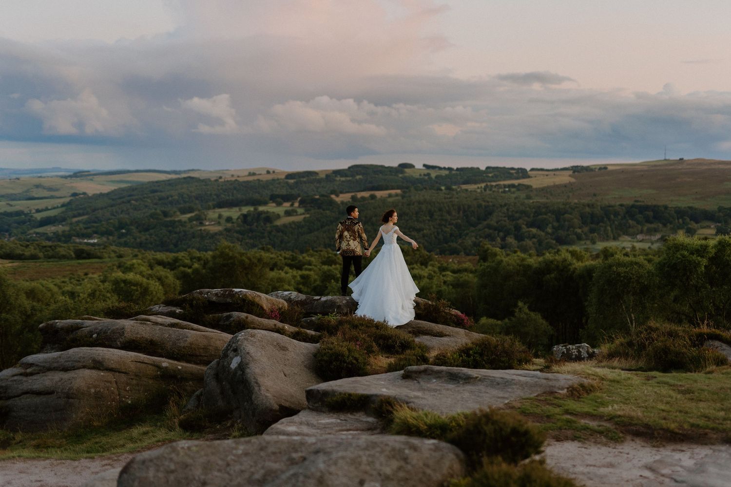 Bride and groom stand on rocky hilltop with sweeping countryside landscape at golden hour.
