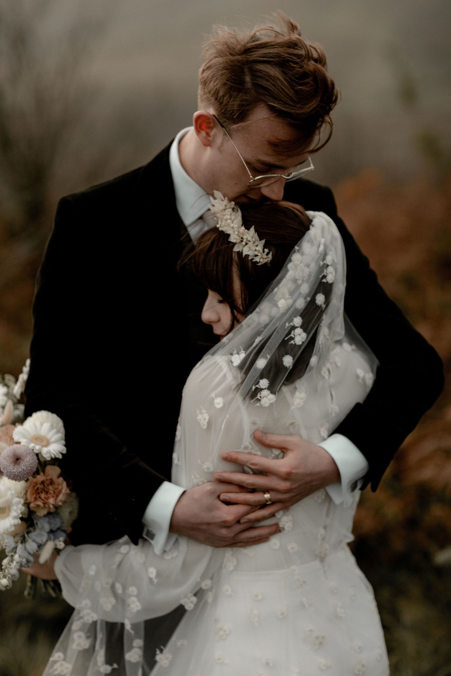 A romantic wedding embrace with the bride in white lace and groom in black suit standing close together outdoors.
