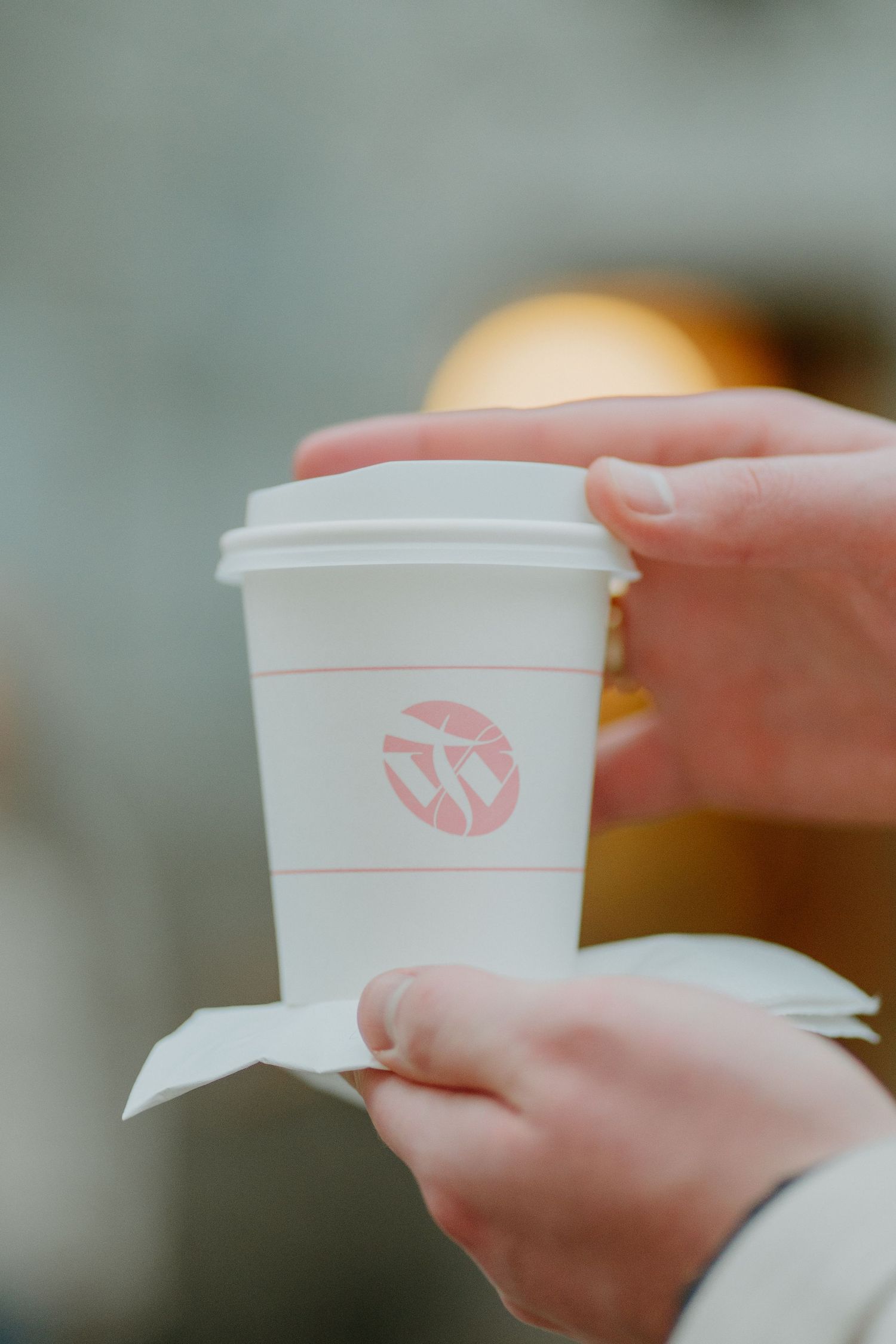 Hands holding a white disposable coffee cup with a pink logo against a blurred background.