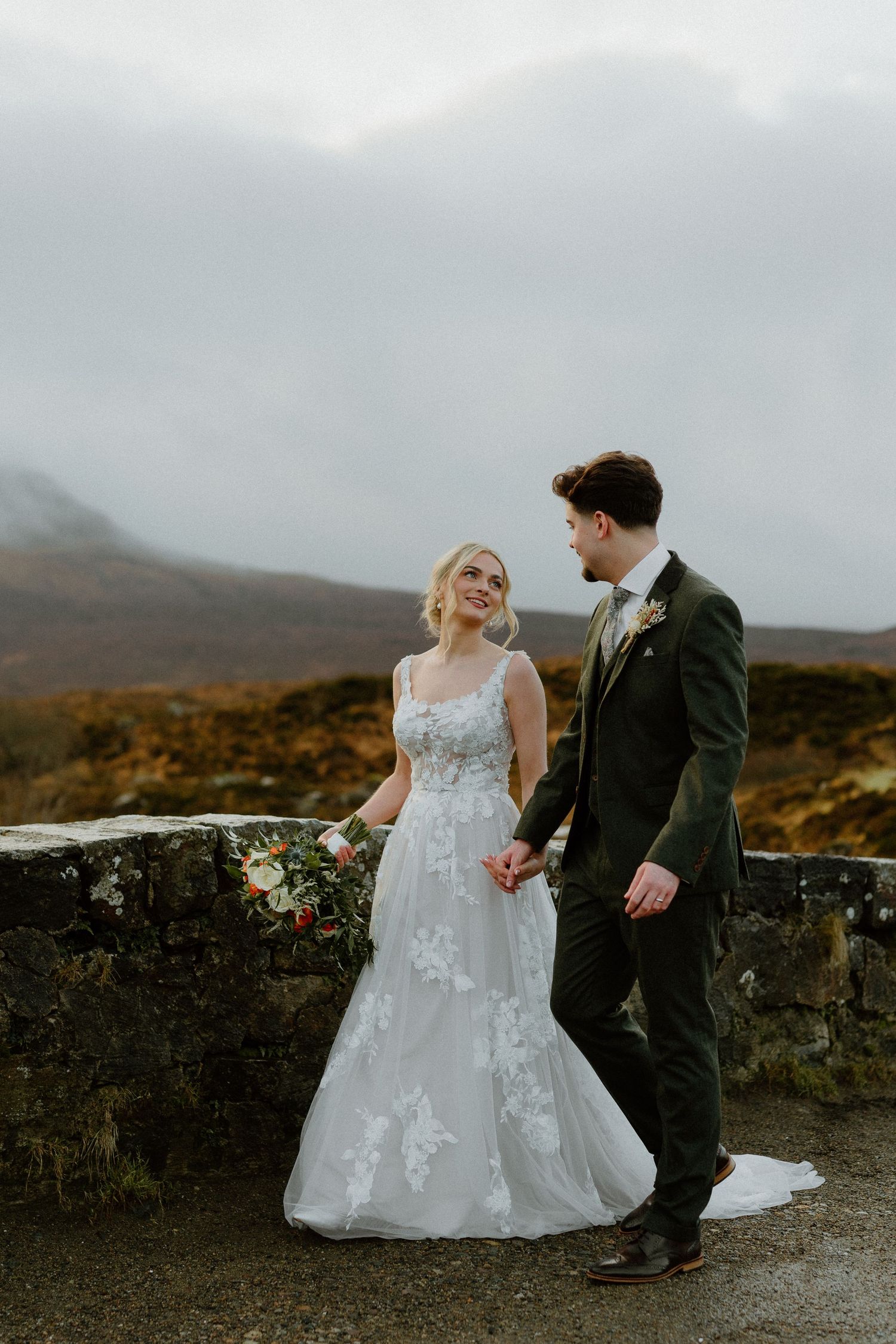 Smiling bride in floral lace gown walks hand-in-hand with groom along stone wall in misty Scottish Highland landscape.