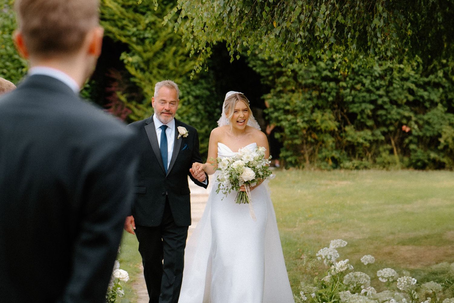 Bride in white gown and veil walks arm-in-arm with father toward groom at outdoor garden ceremony.