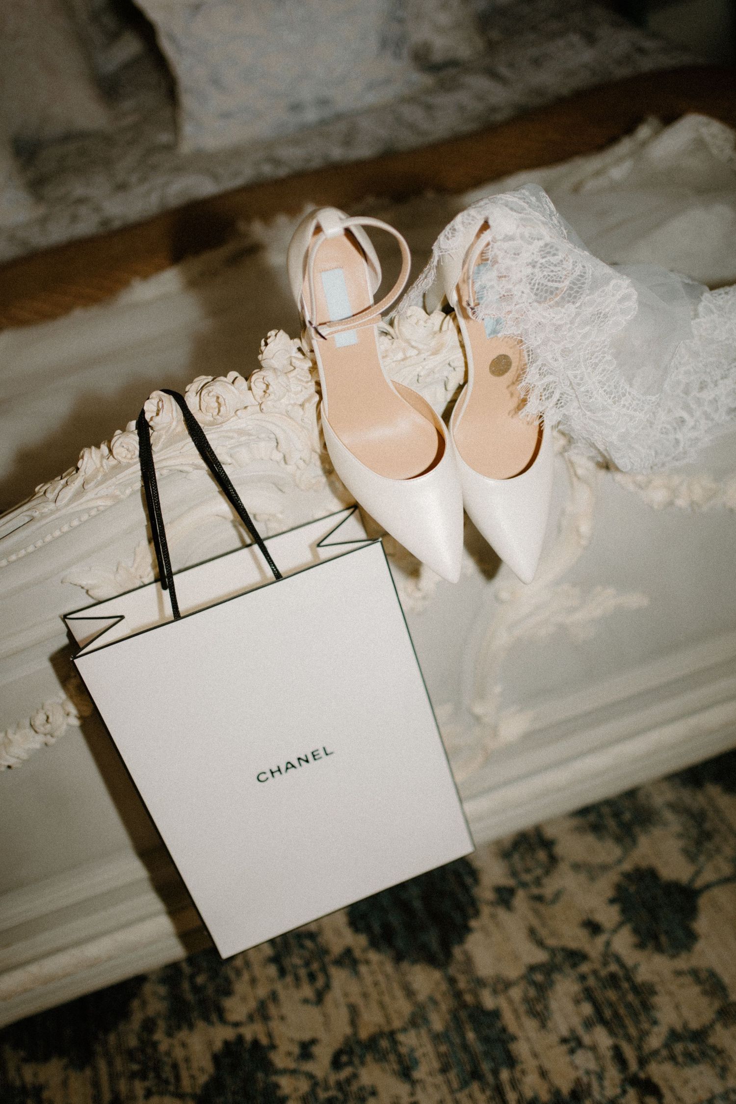 Chanel shopping bag with white bridal heels and lace veil displayed on an ornate white surface.